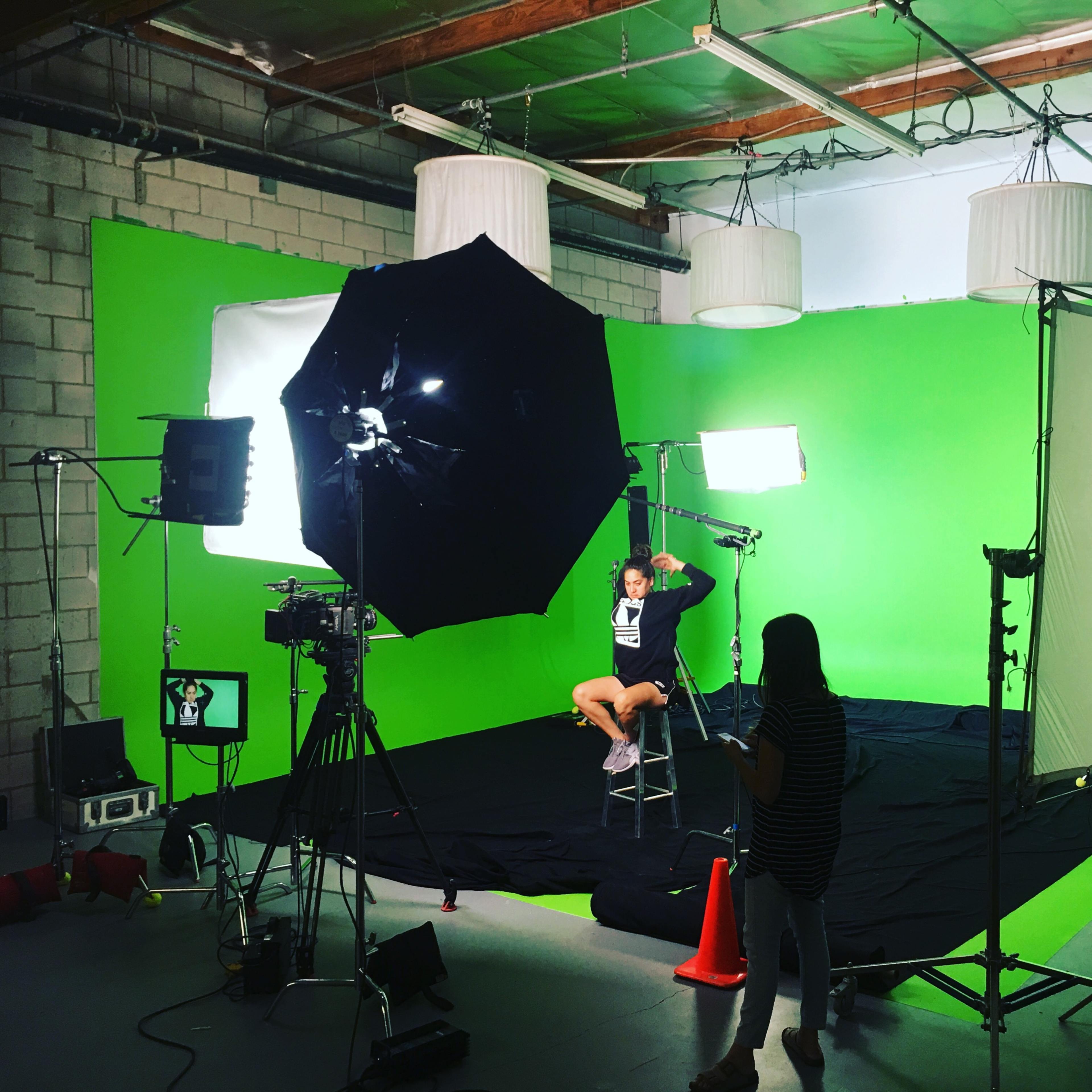 A person sits on a stool in front of a green screen surrounded by various cameras and lighting equipment in a studio setting.