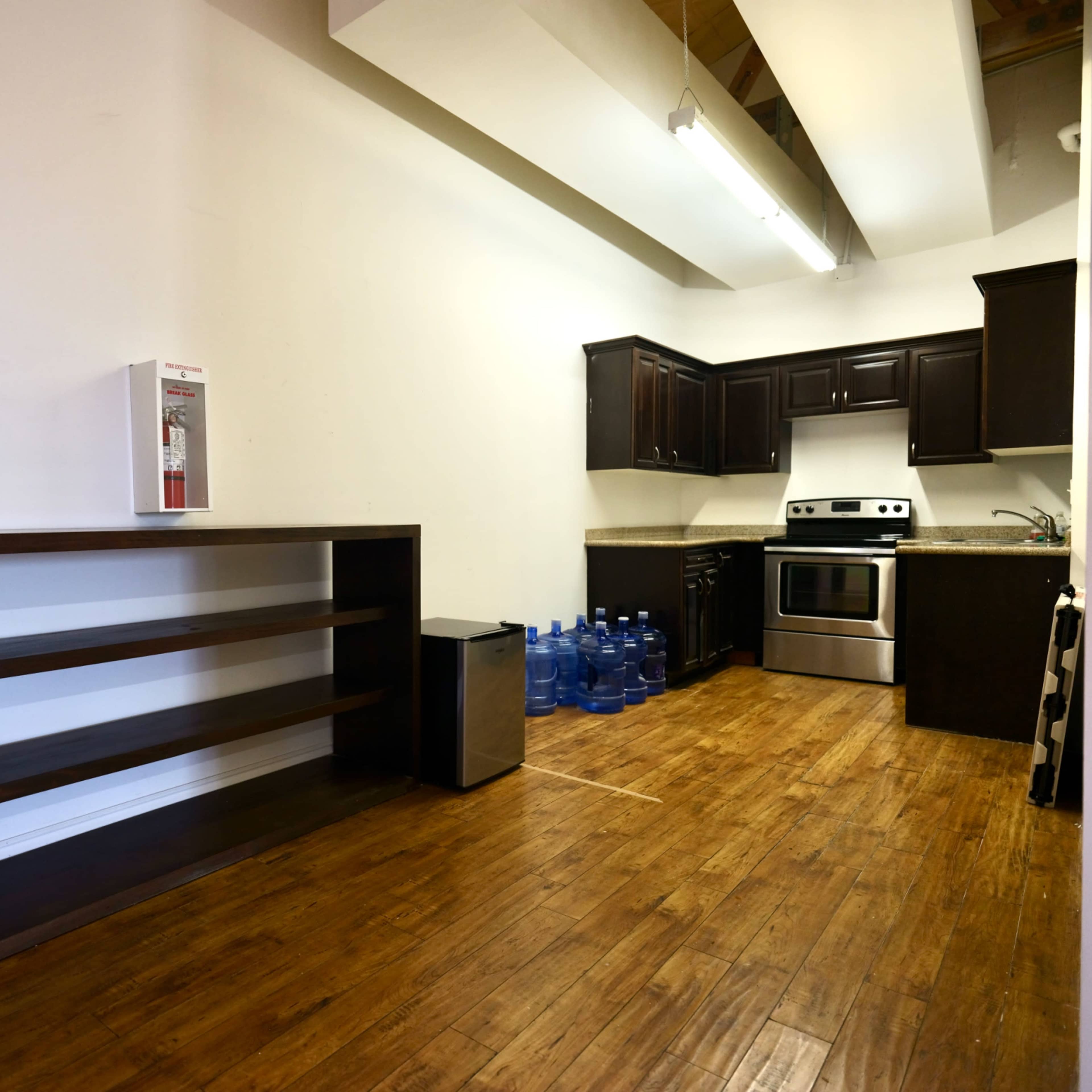 A kitchen area with dark wooden cabinetry, a stainless steel stove, and a small refrigerator, alongside a shelf and multiple water bottles on the floor.