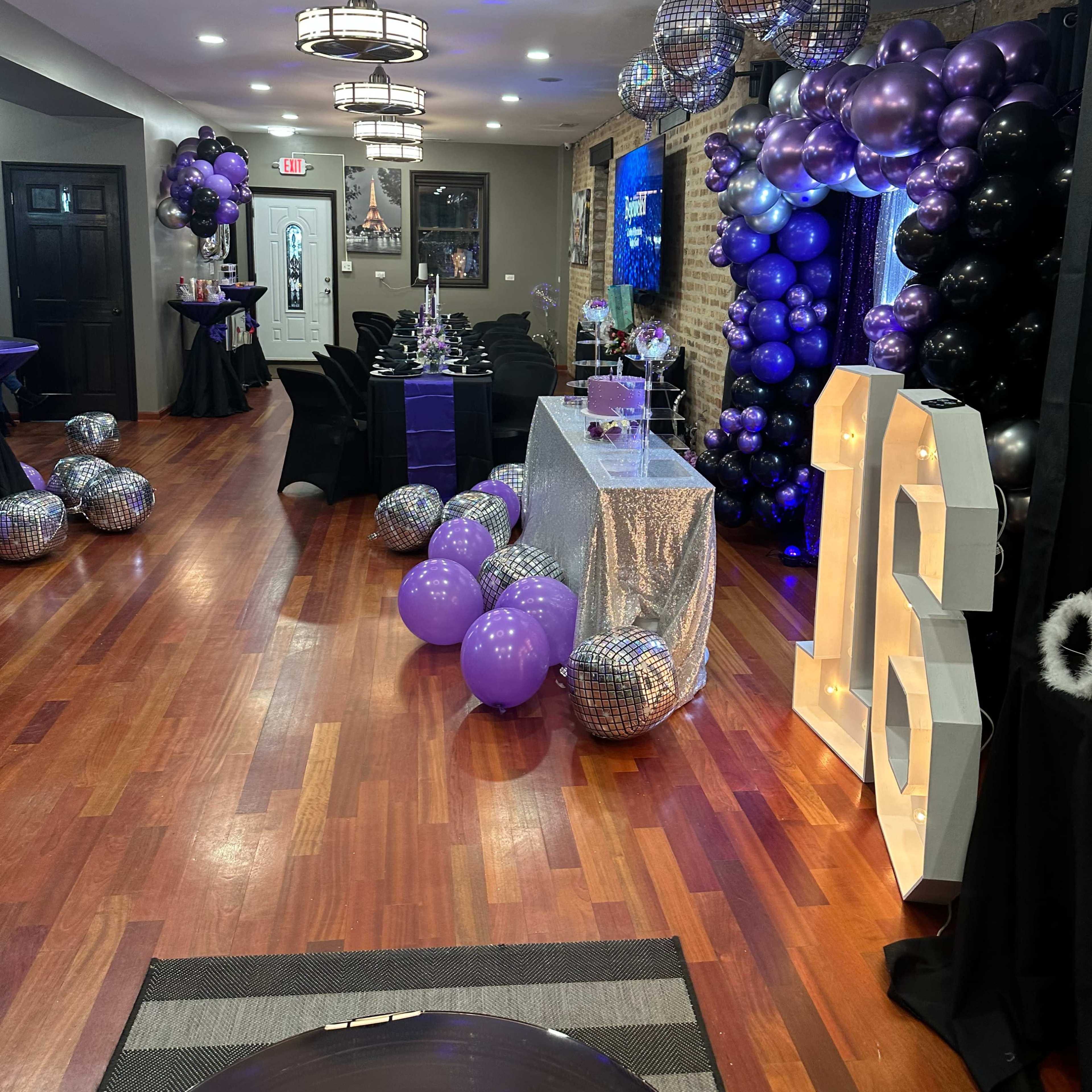 A decorated event space featuring a sparkling silver table, black and purple balloons, and a large illuminated "16" sign.