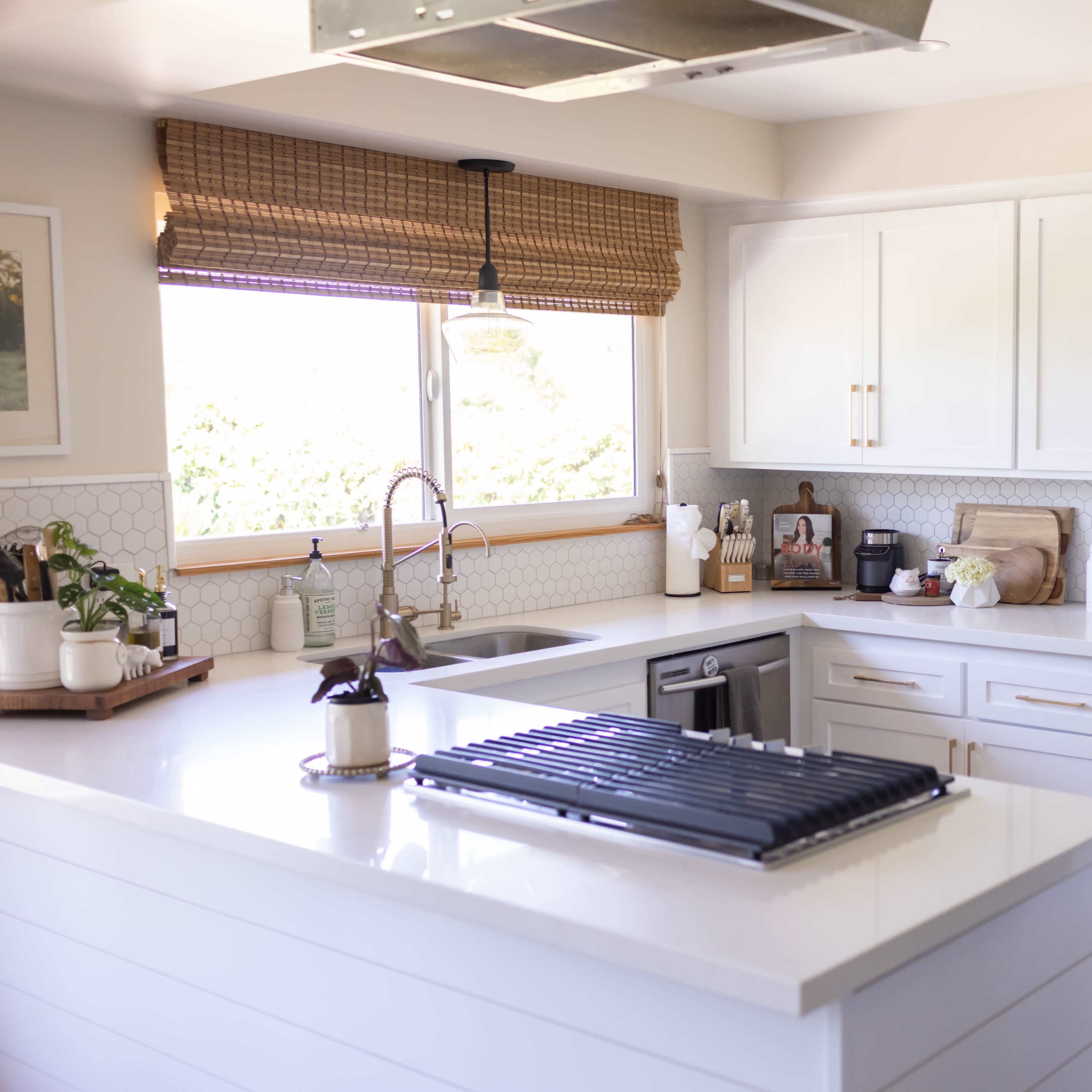 A modern kitchen features white cabinetry, a large island with a cooktop, and a window with natural light filtering in through woven shades.