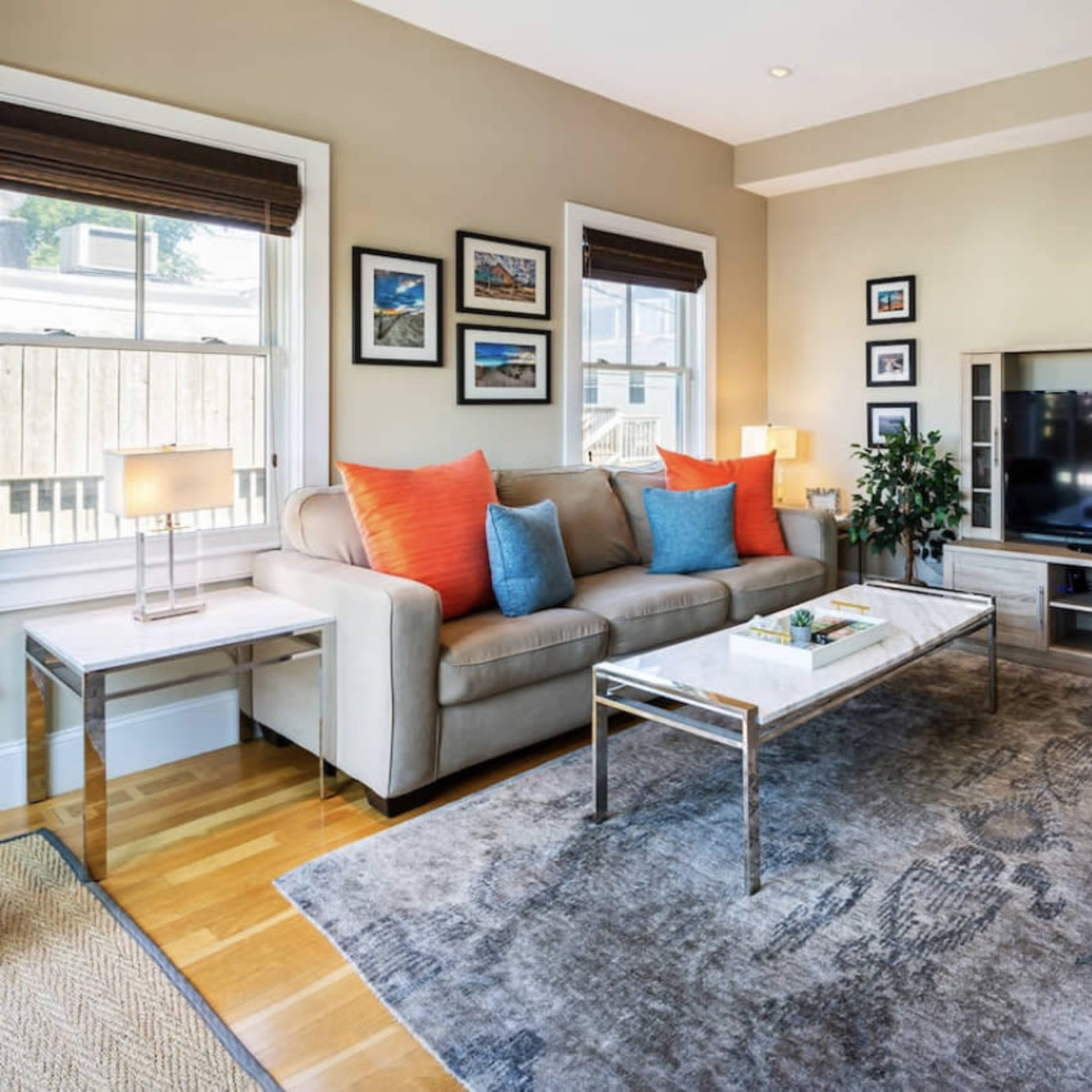 A living room features a light gray sofa with orange cushions, a coffee table, a television unit, and a patterned area rug, all arranged around a wooden floor.