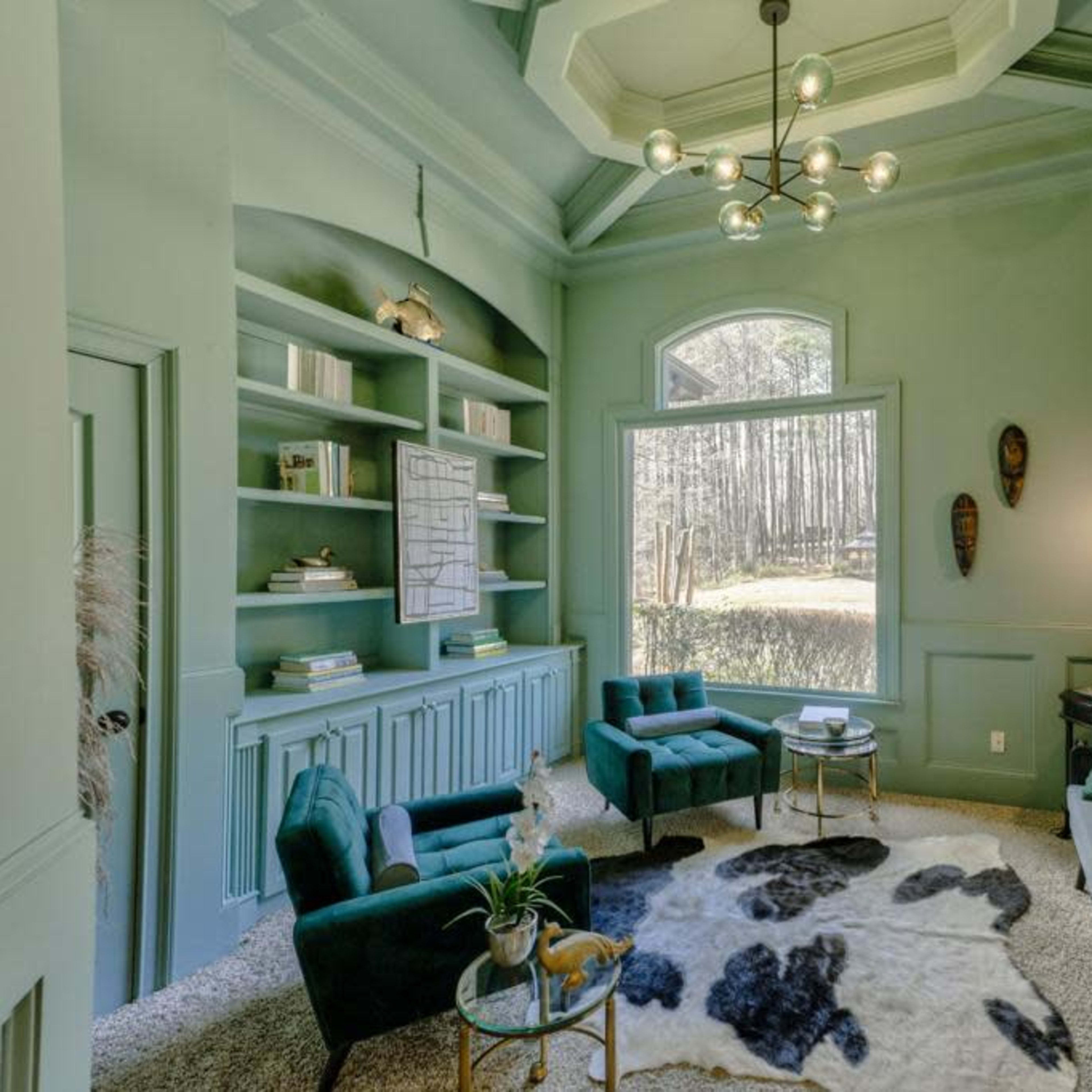 A cozy living space features teal walls, a plush carpet, a large window overlooking trees, and various seating arrangements.