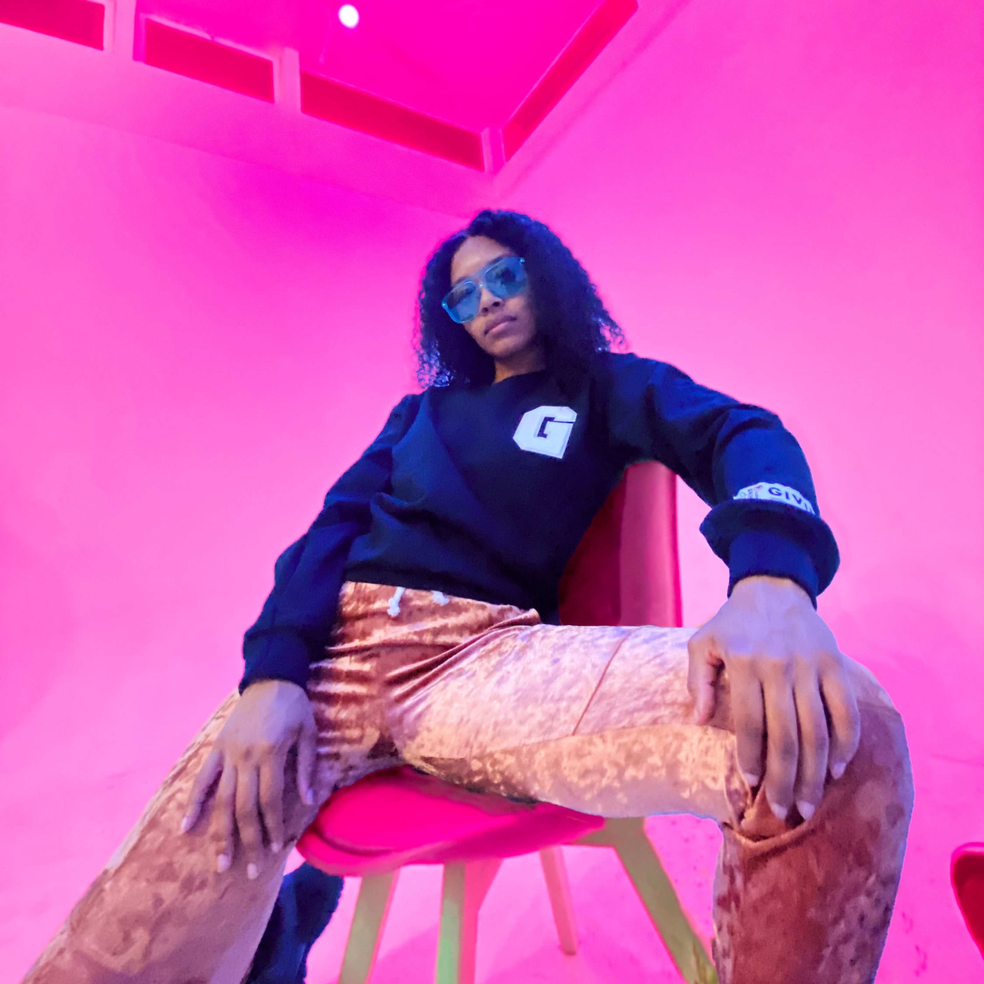 A person with curly hair poses casually in a black sweatshirt and shiny gold pants on a pink chair against a vibrant pink background.
