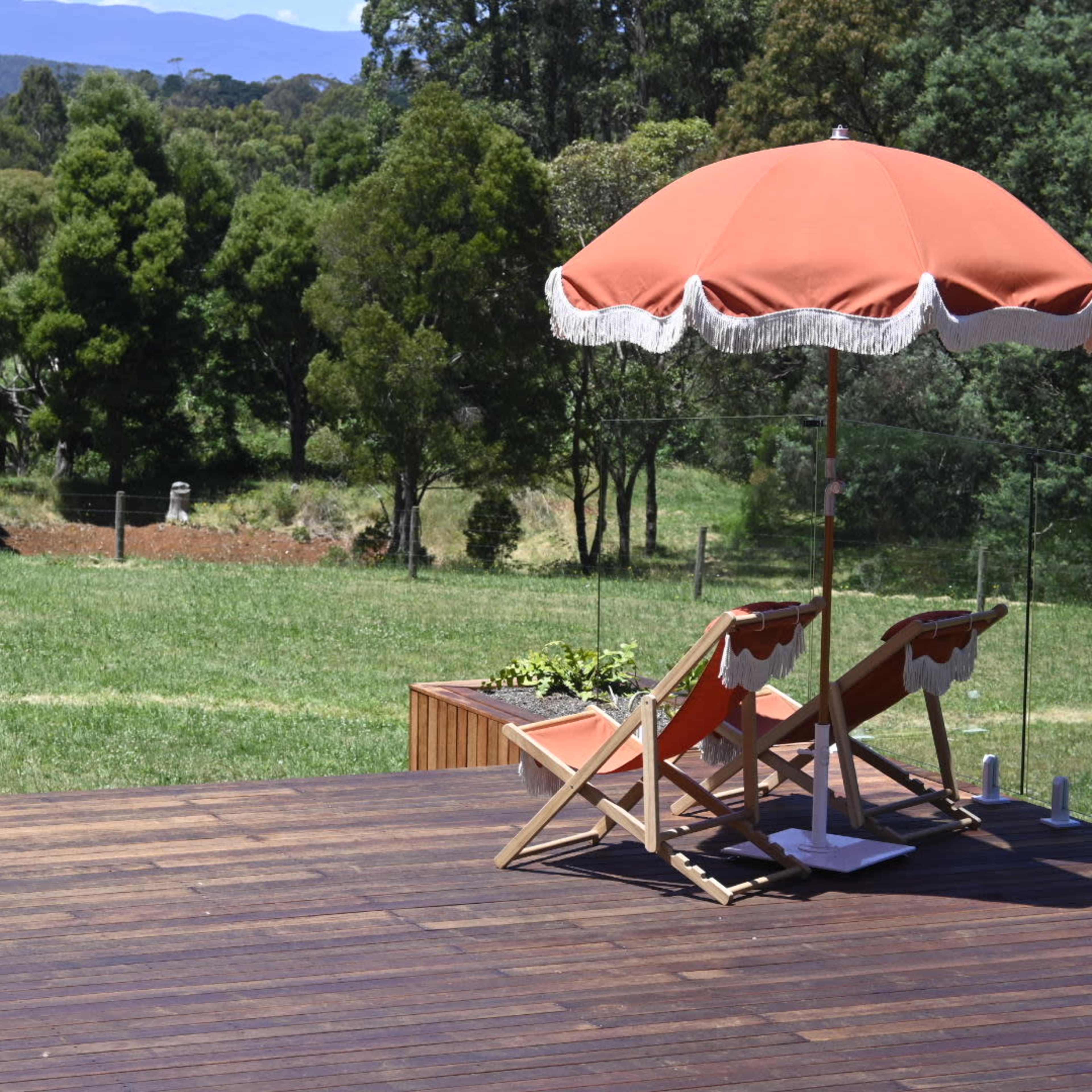 A wooden deck features two lounge chairs under a red umbrella, overlooking a grassy area with trees and mountains in the background.