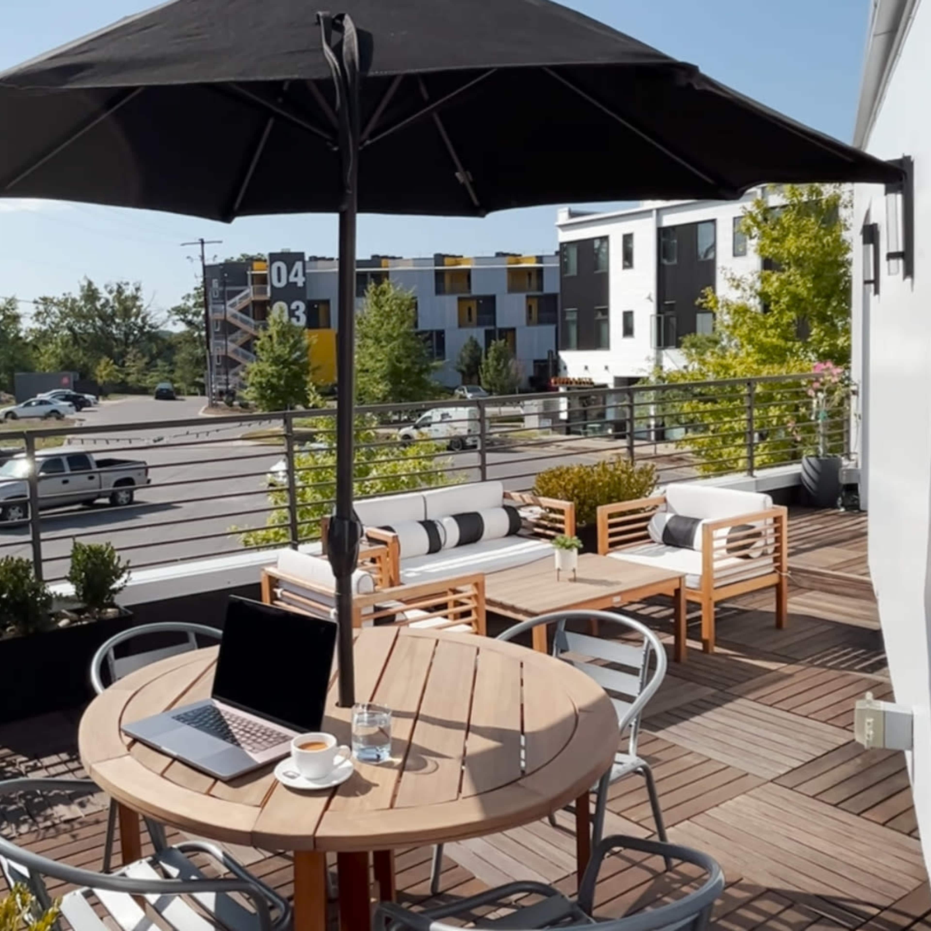 A laptop and a coffee cup sit on a round table beneath a large umbrella on a balcony overlooking a street with passing cars and modern buildings.