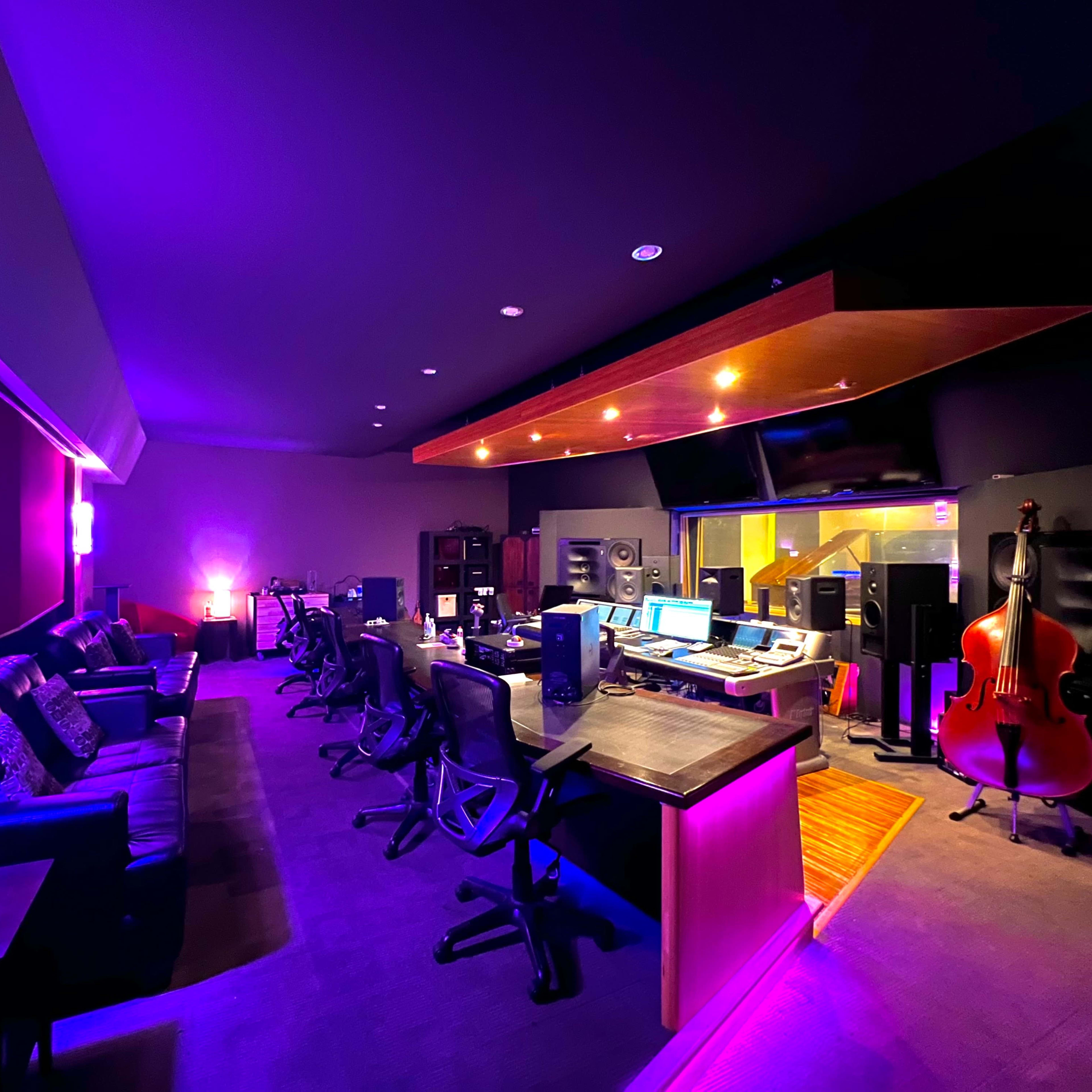A dimly lit music studio featuring a control area with audio equipment, multiple chairs, and a large double bass in the corner.