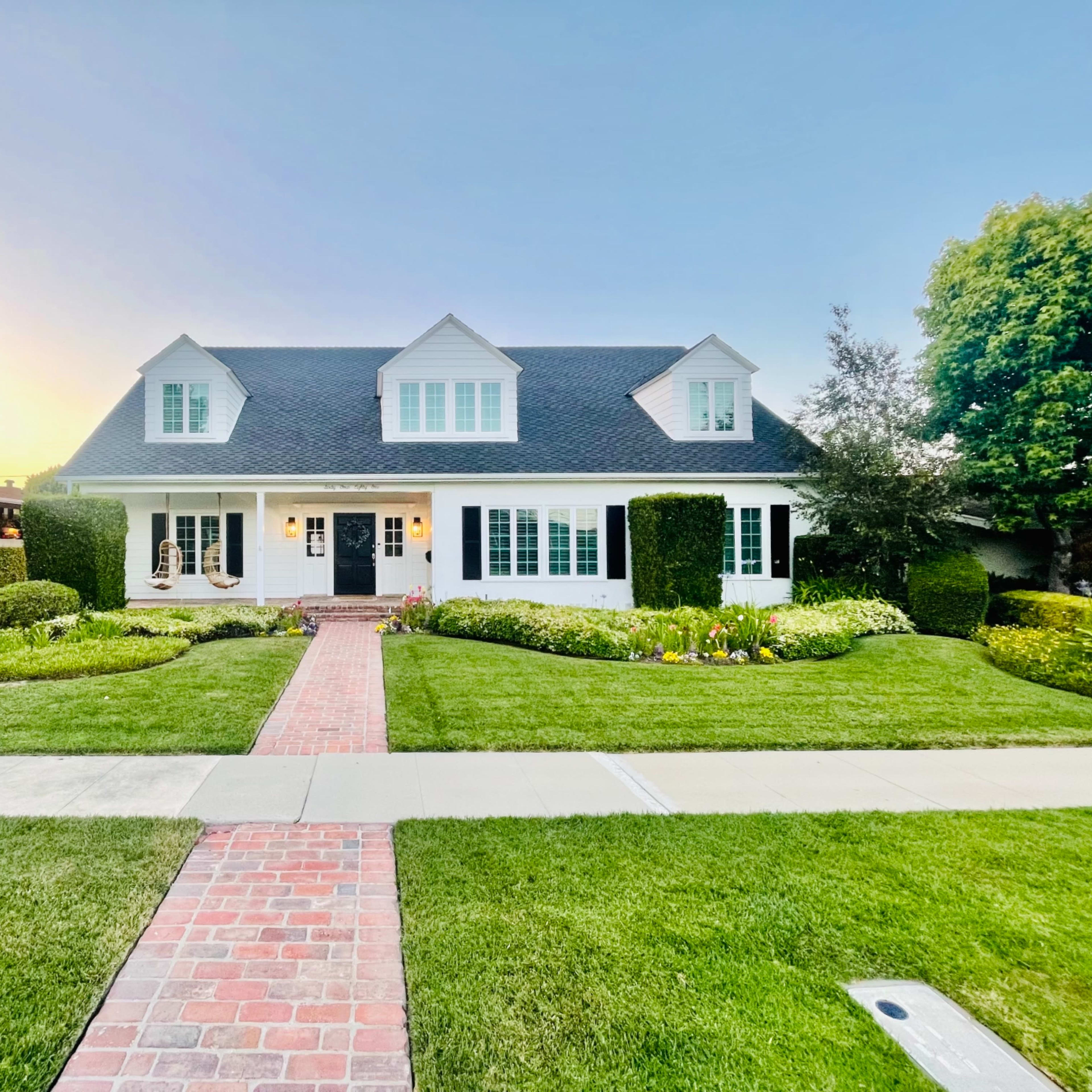 A two-story white house with a dark roof and neatly trimmed hedges is surrounded by a manicured lawn and colorful flower beds.
