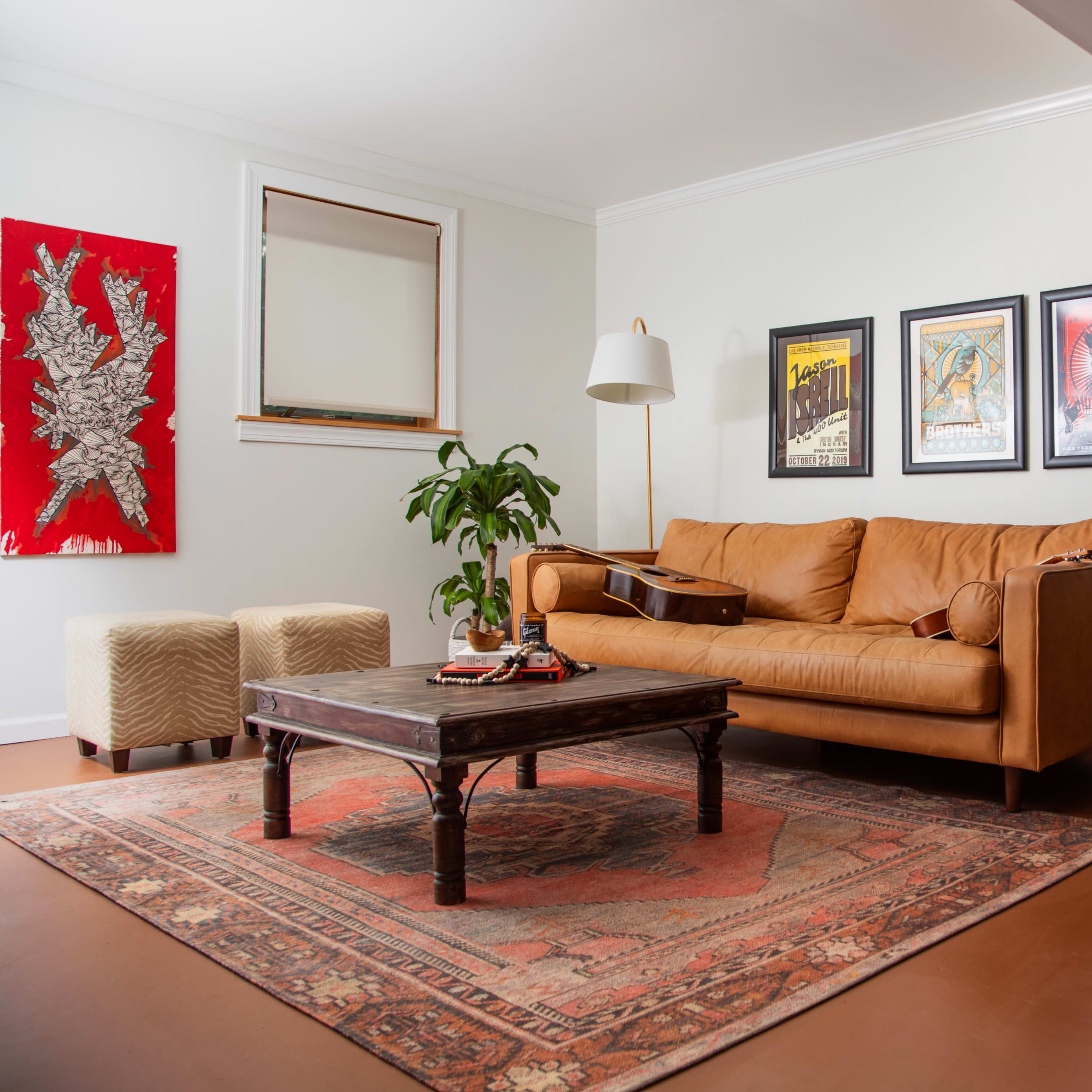 A cozy living room features a brown leather couch, a coffee table, framed artwork on the walls, a guitar resting on the couch, and an amplifier in the corner.