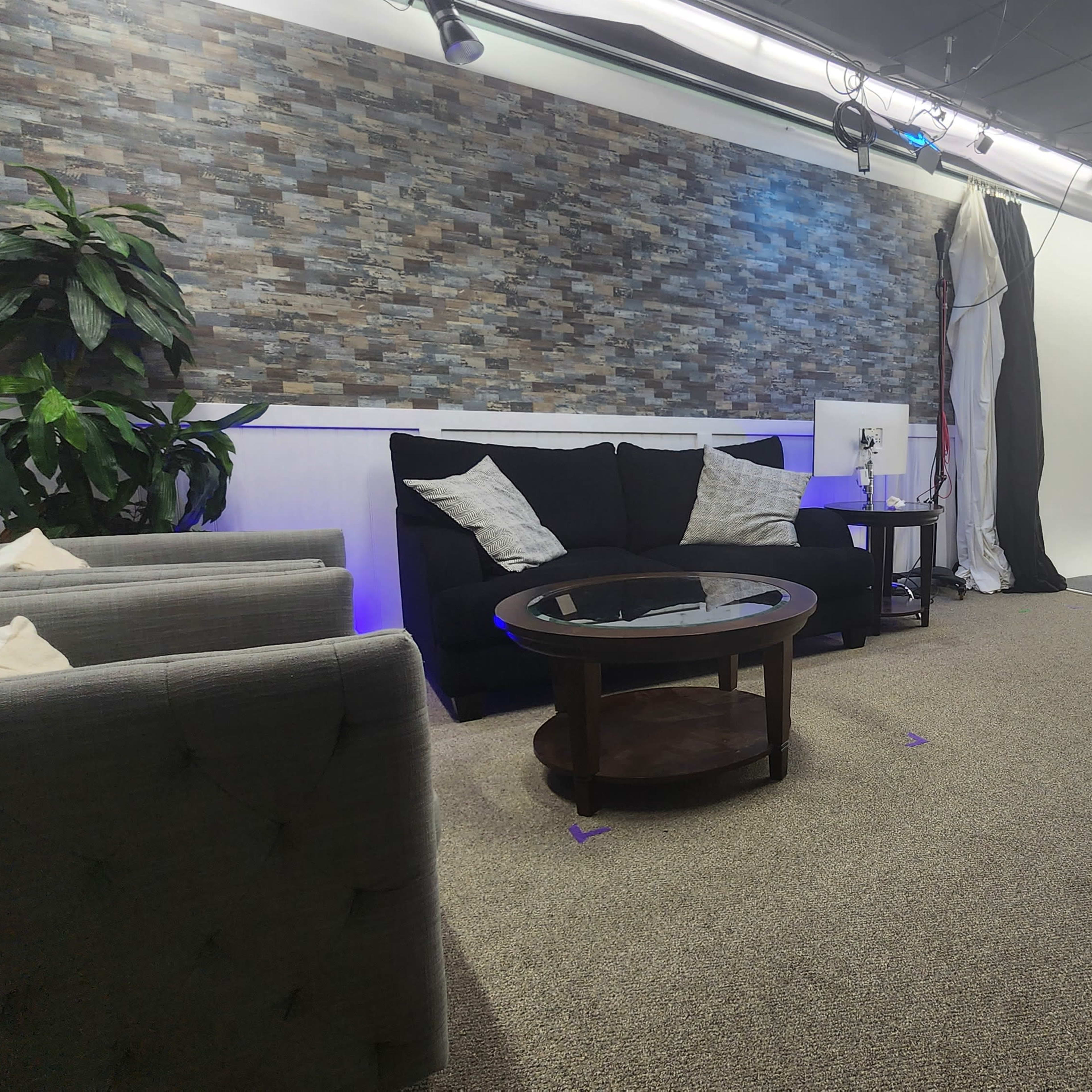 A spacious studio with a carpeted floor features a black couch and a round table, illuminated by soft lighting against a stone-patterned wall.