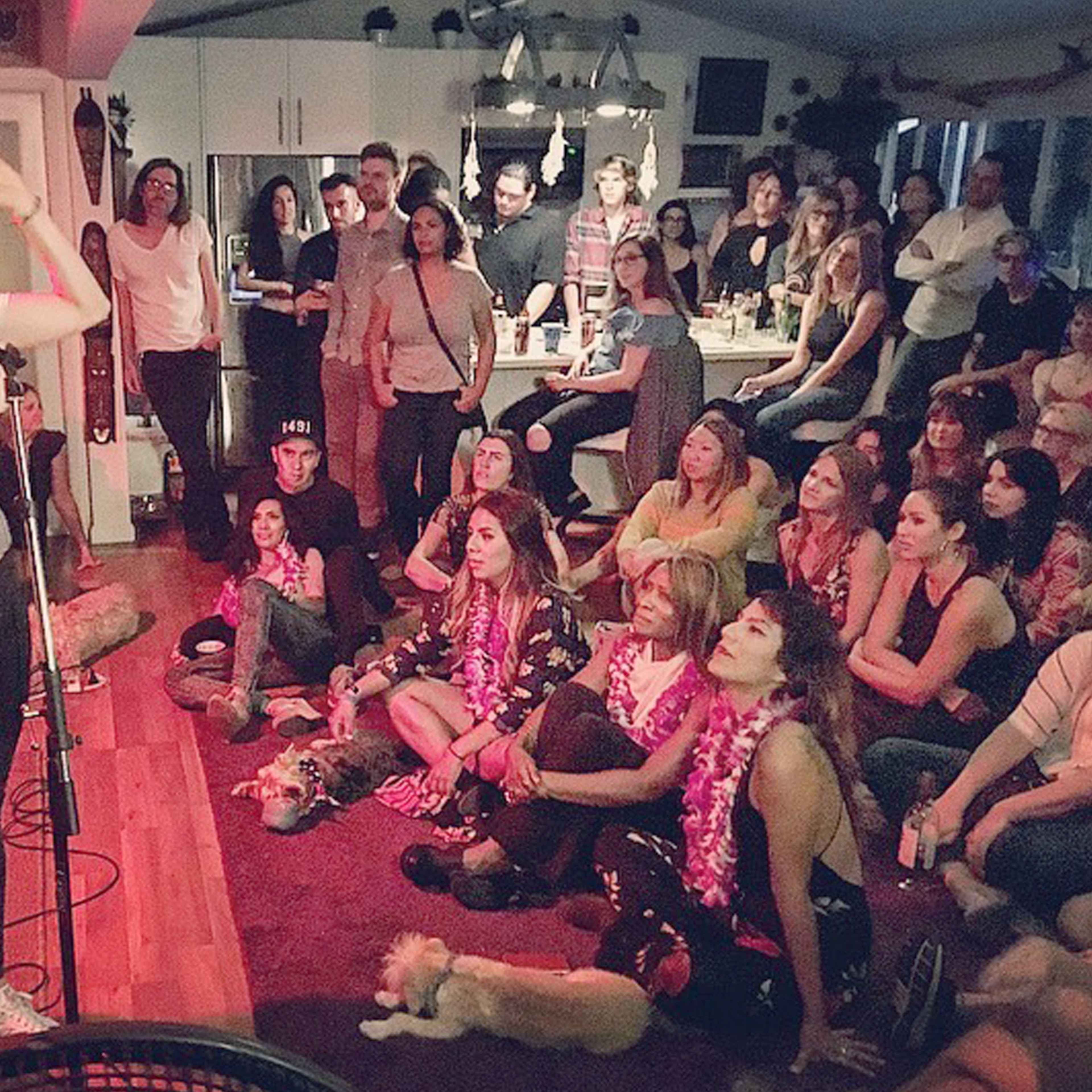 A performer stands on a microphone in front of an audience seated on the floor, while others watch from the back of a lively venue.