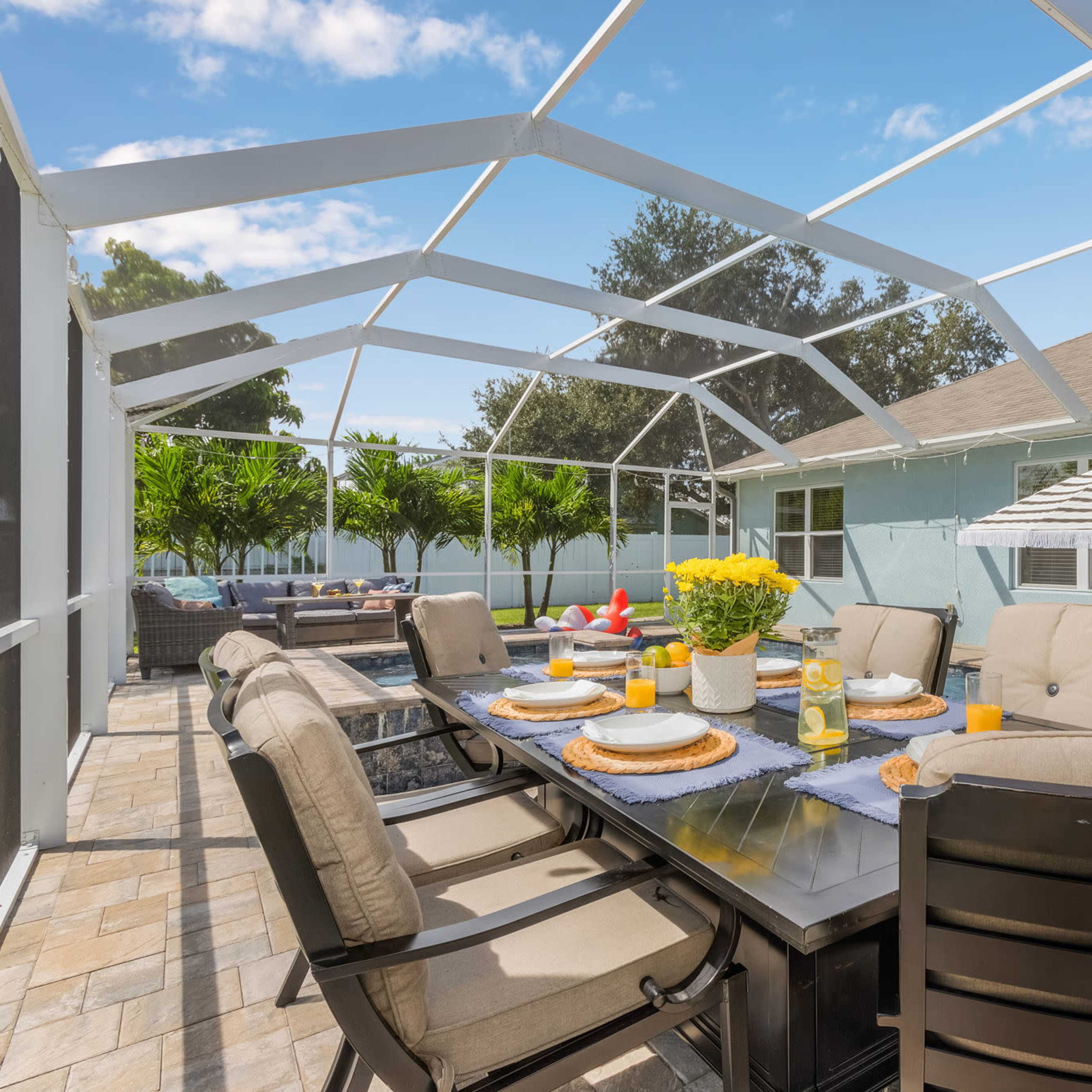 A screened patio features a dining table set with plates and drinks, surrounded by cushioned chairs and tropical plants, with a view of a pool.
