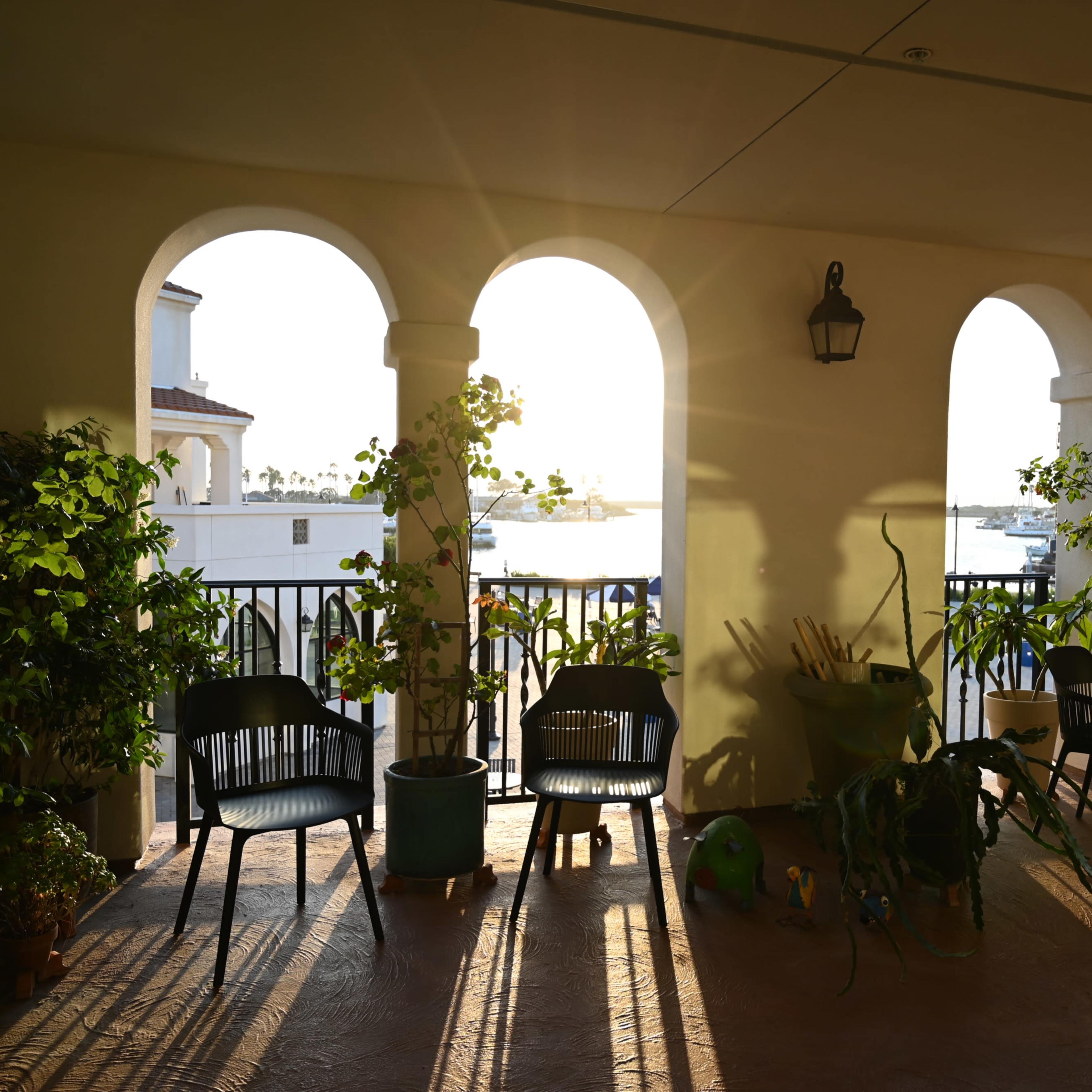 The scene shows a brightly lit patio with arches, potted plants, and black chairs, overlooking water in the background.