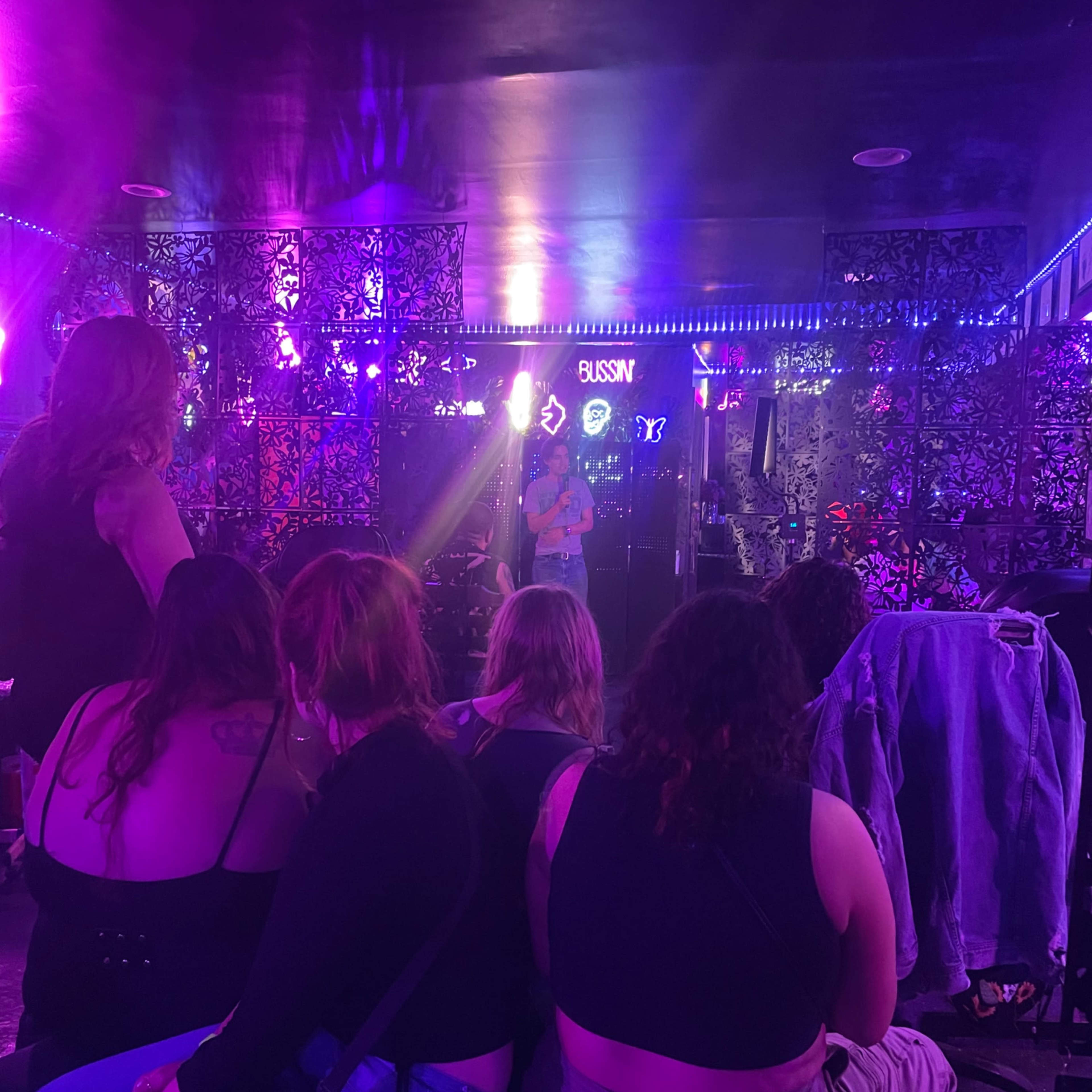 A performer stands on a stage in a dimly lit venue surrounded by patrons seated on couches.