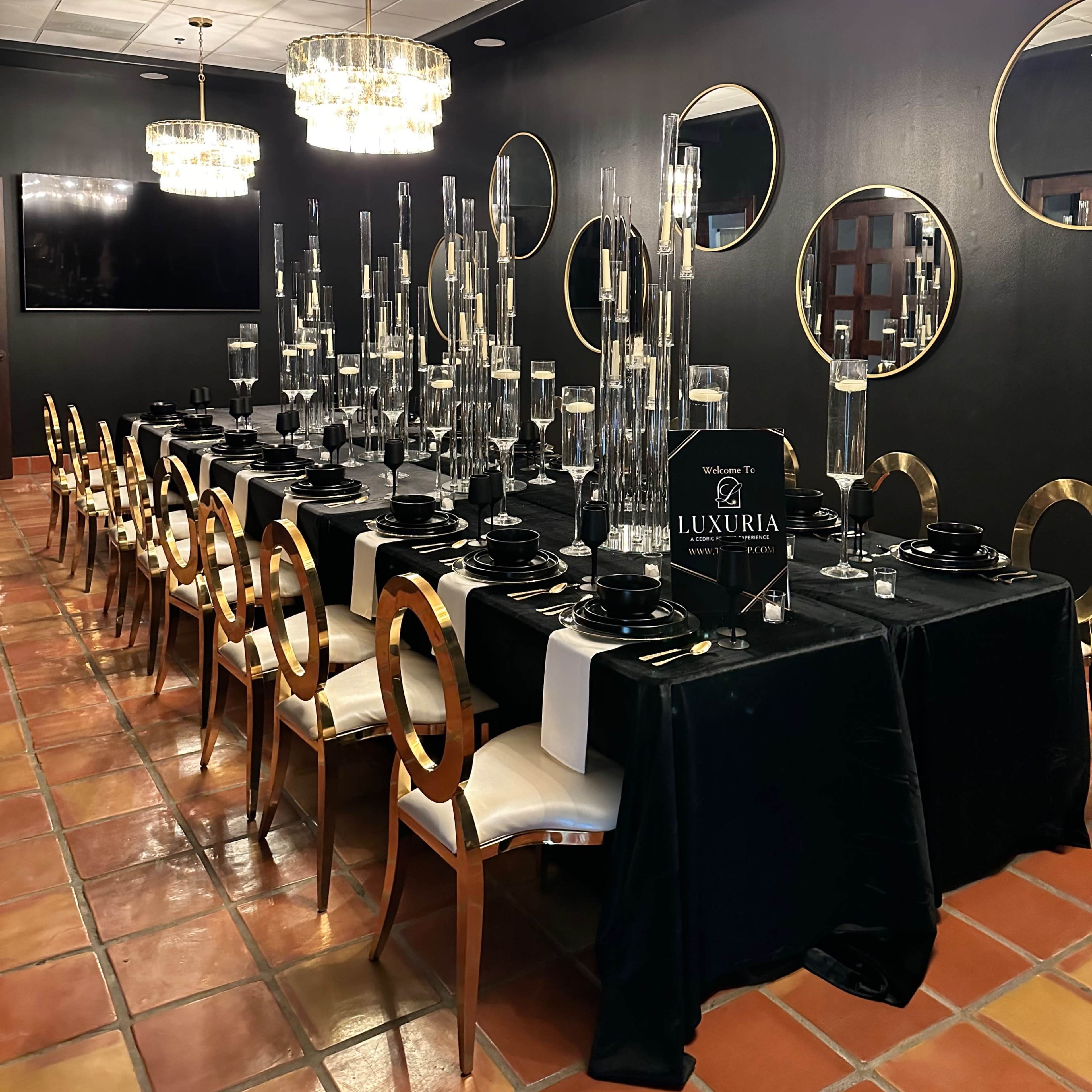 A long dining table is elegantly set in a dark room, featuring tall glass centerpieces and surrounded by gold-accented chairs.