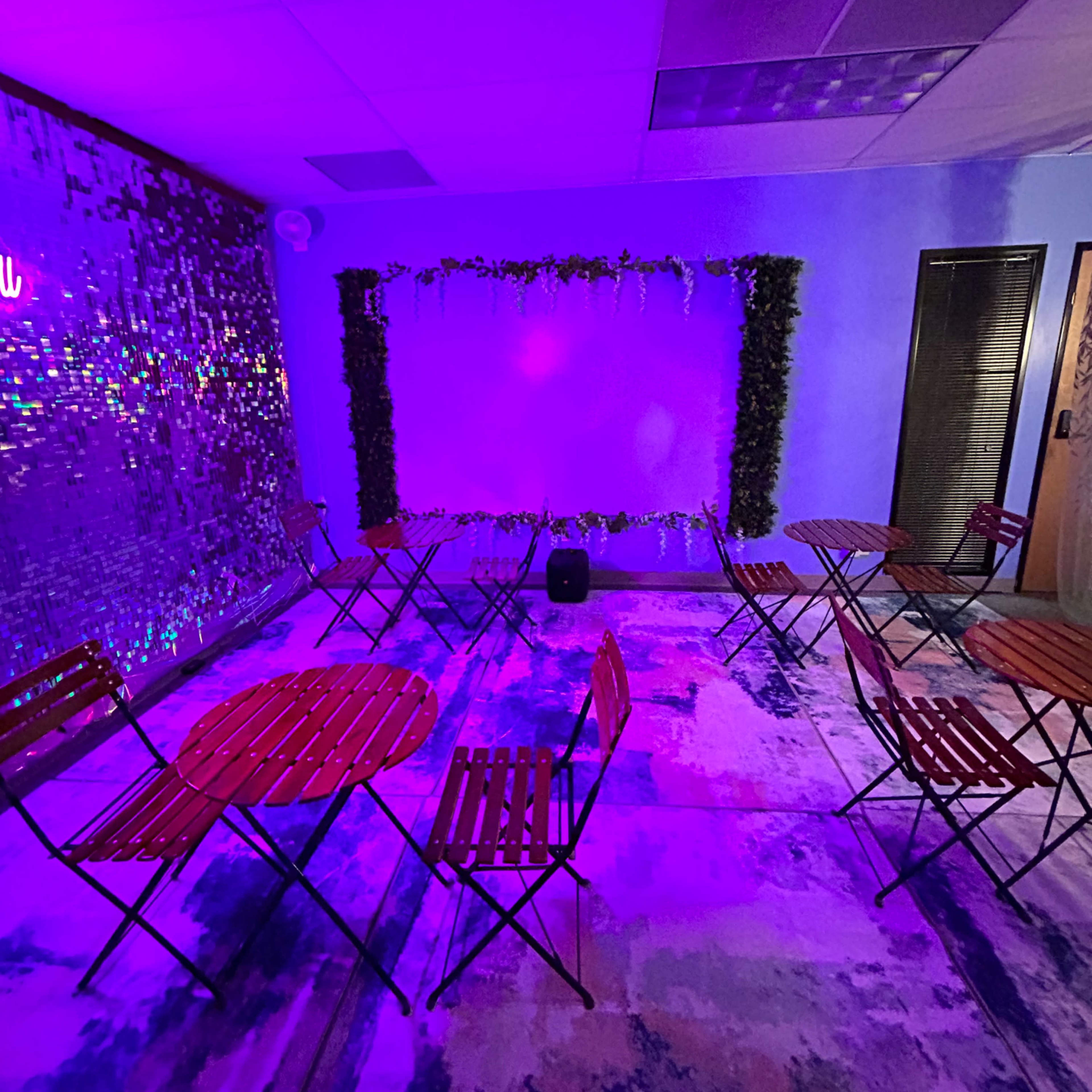 A room with several round wooden tables and chairs arranged around a purple-lit wall adorned with greenery.