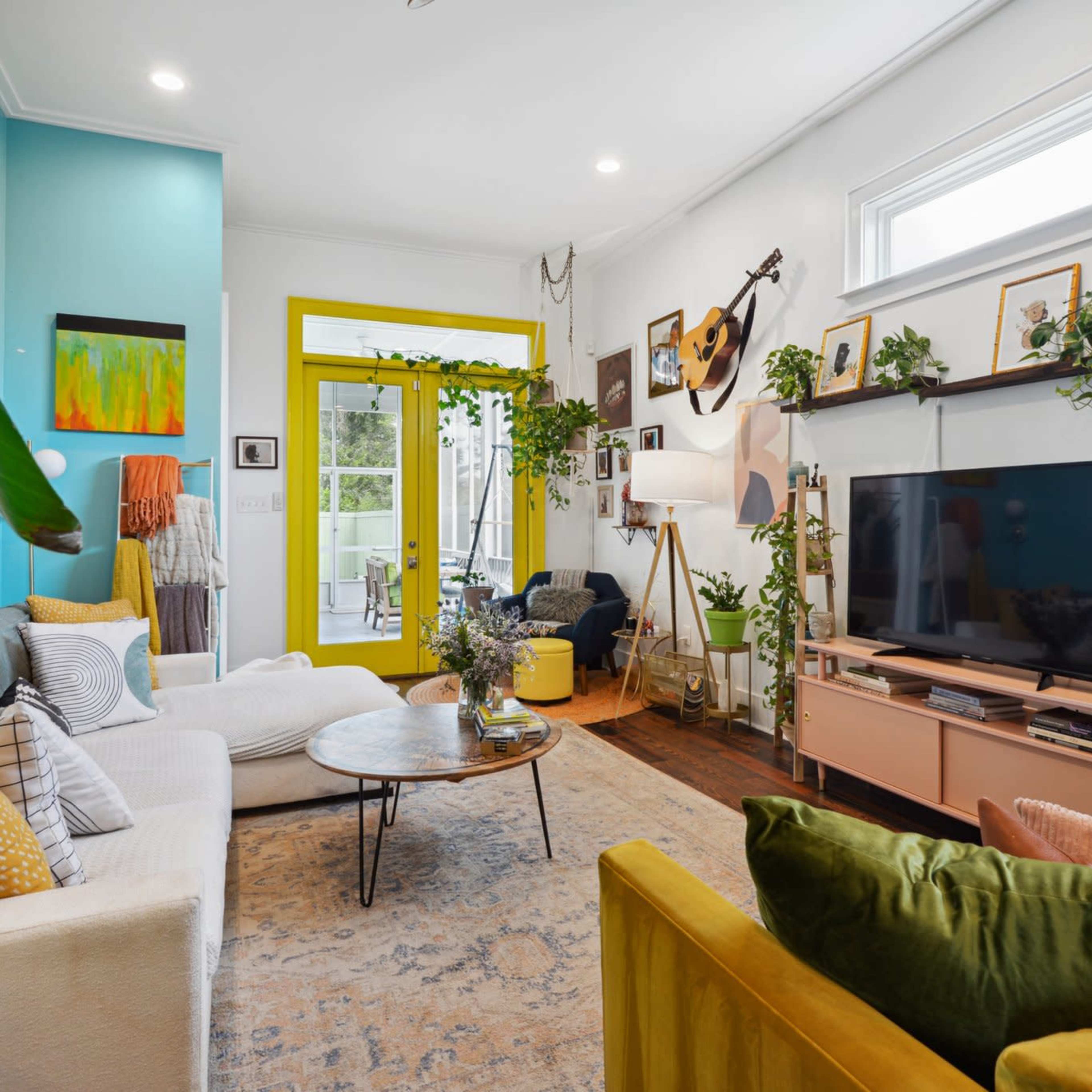 A colorful living room features two sofas, a round coffee table, a television on a light wood console, and plants decorating the walls and furniture.
