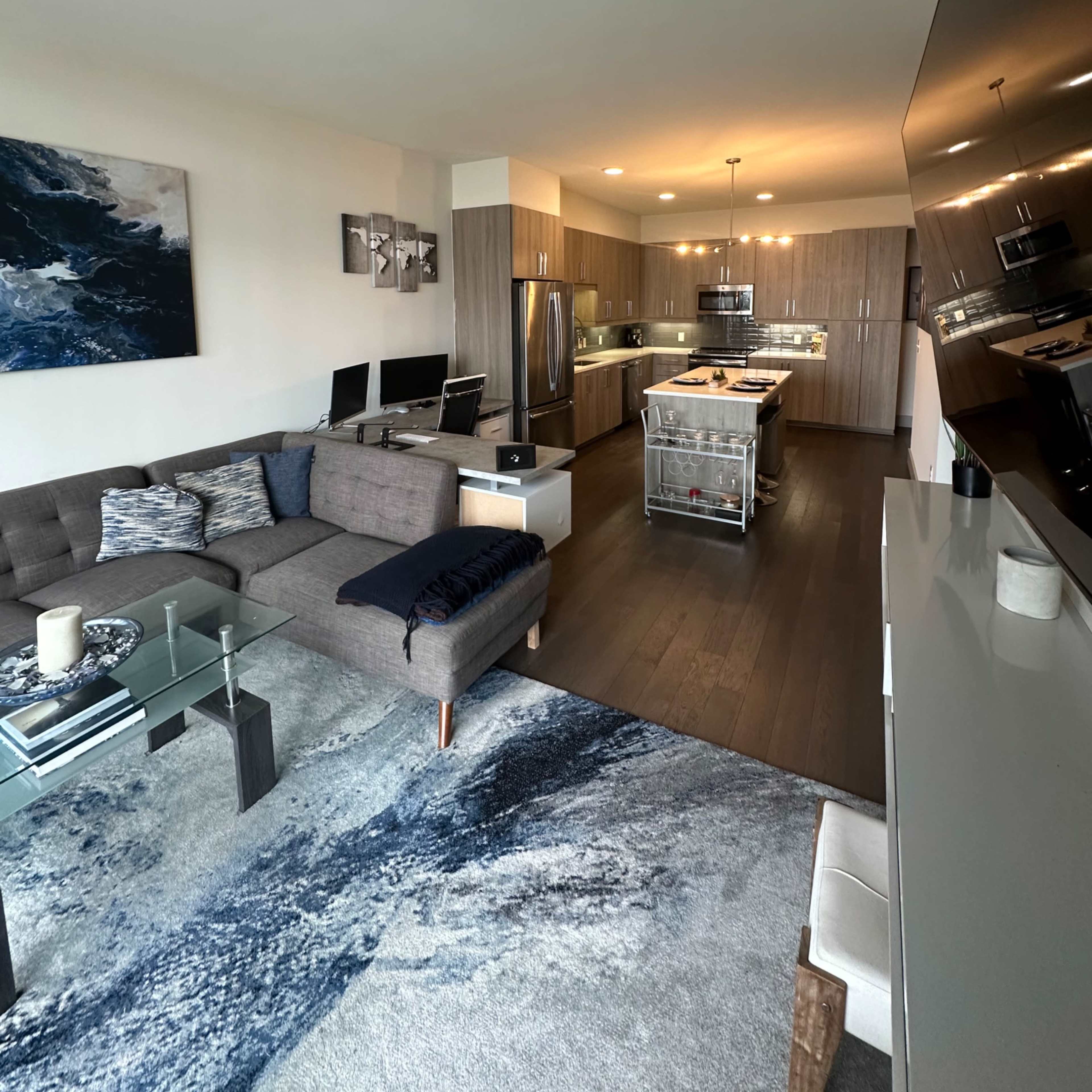 A modern, open-concept living room and kitchen area featuring a sectional sofa, a glass coffee table, and a central island in the kitchen.