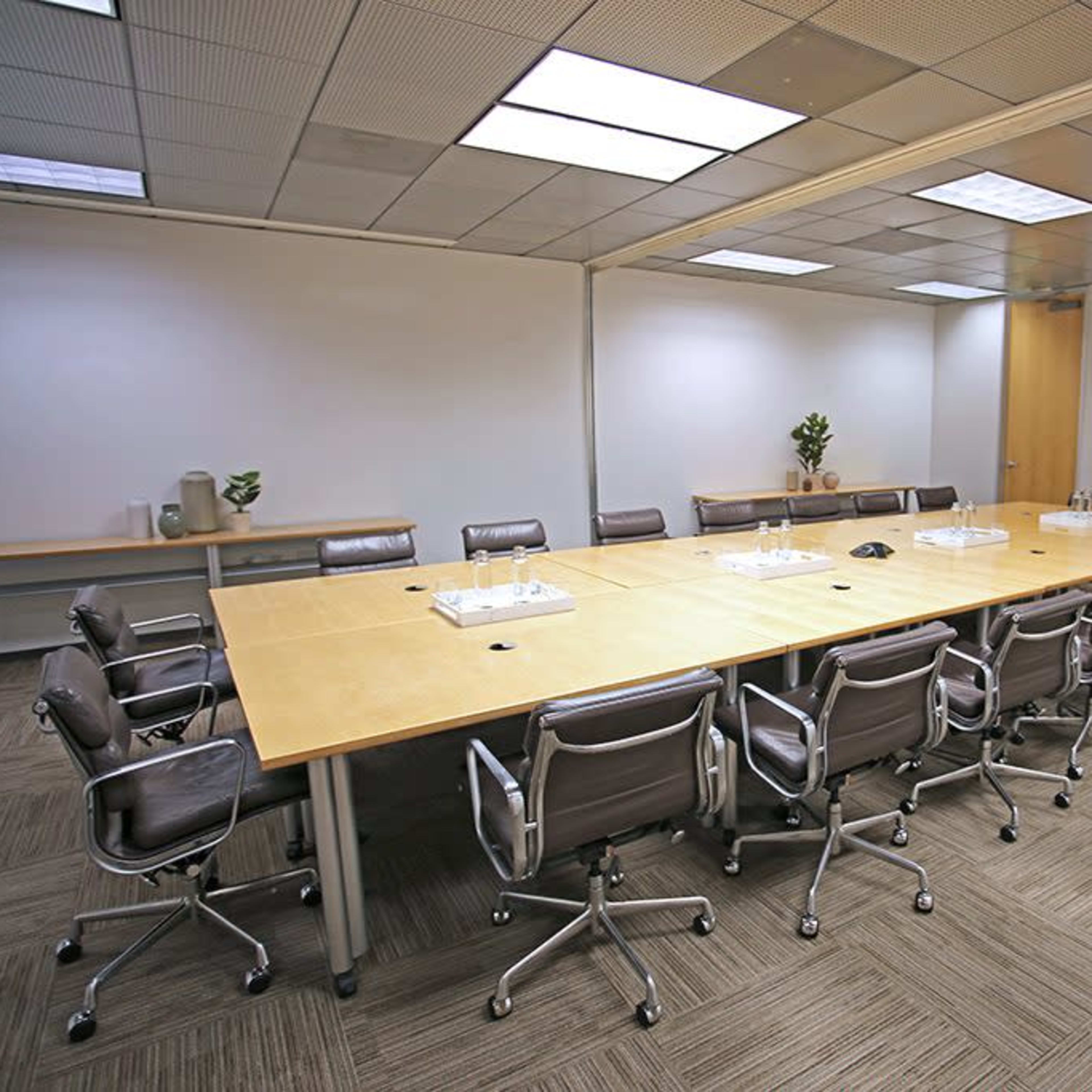 A spacious conference room with a large wooden table surrounded by several gray office chairs, and a plant in a corner.