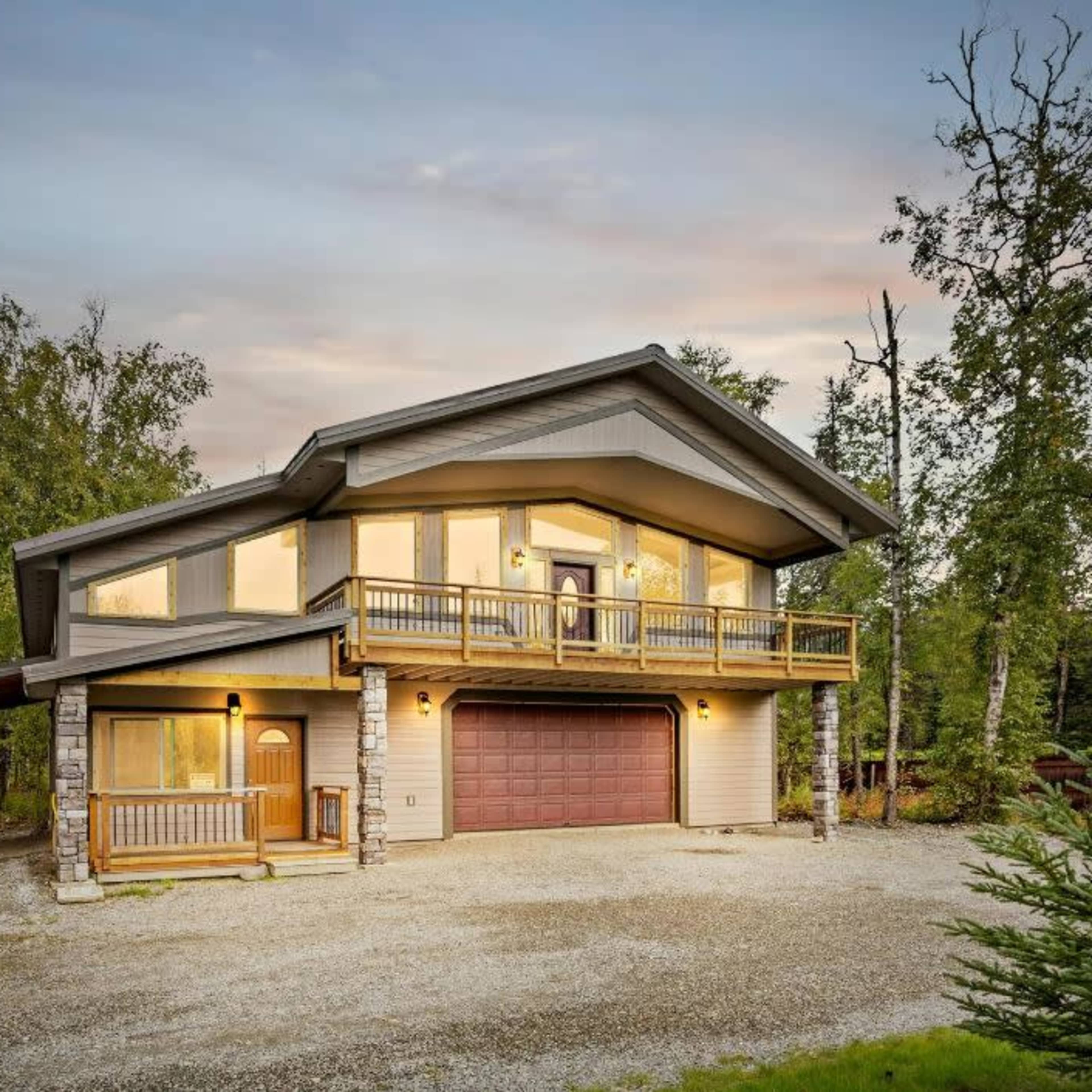 A modern two-story house with a large front deck and a garage is surrounded by trees and gravel driveway.