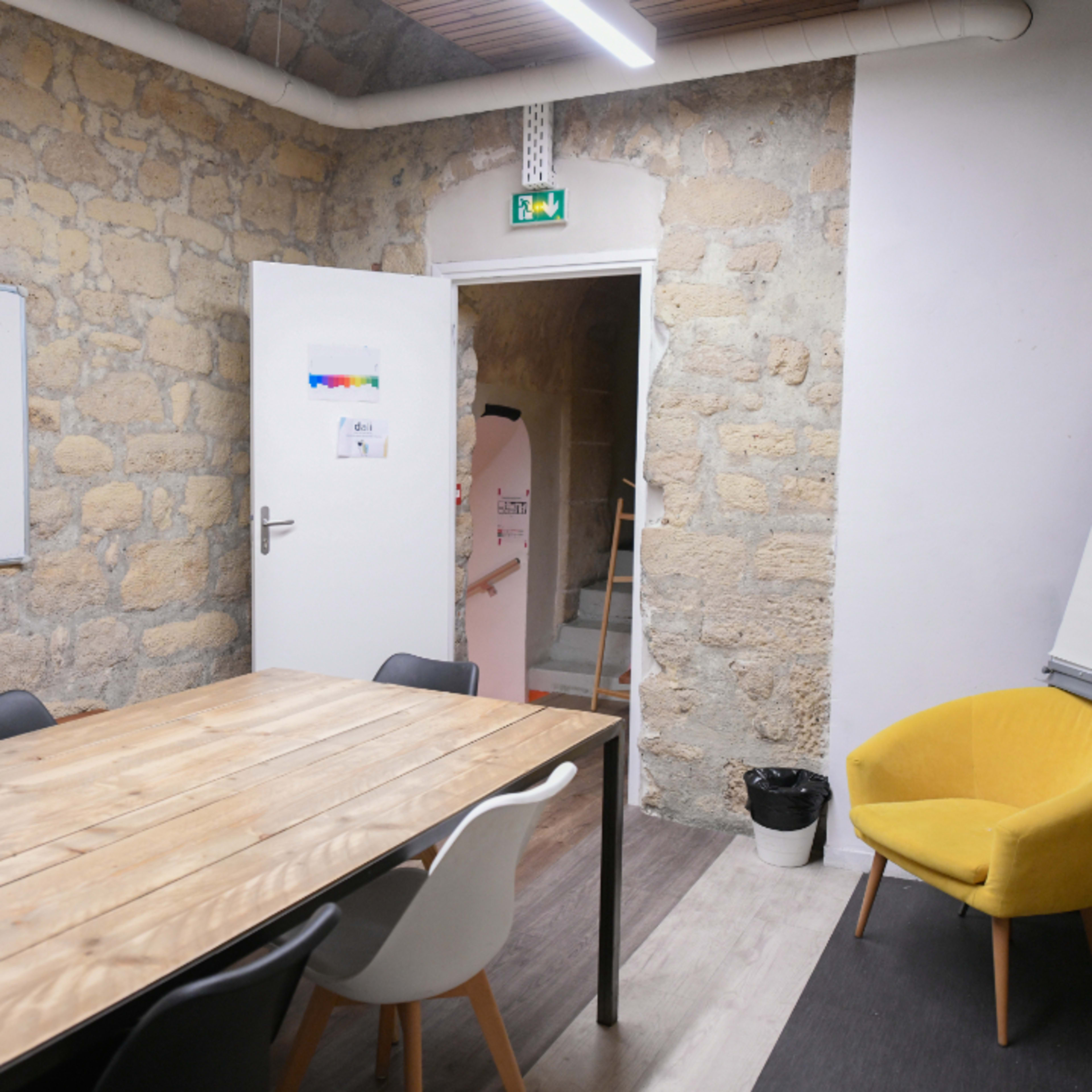 A meeting room with a wooden table and chairs, a yellow armchair in the corner, and a doorway leading to a staircase.