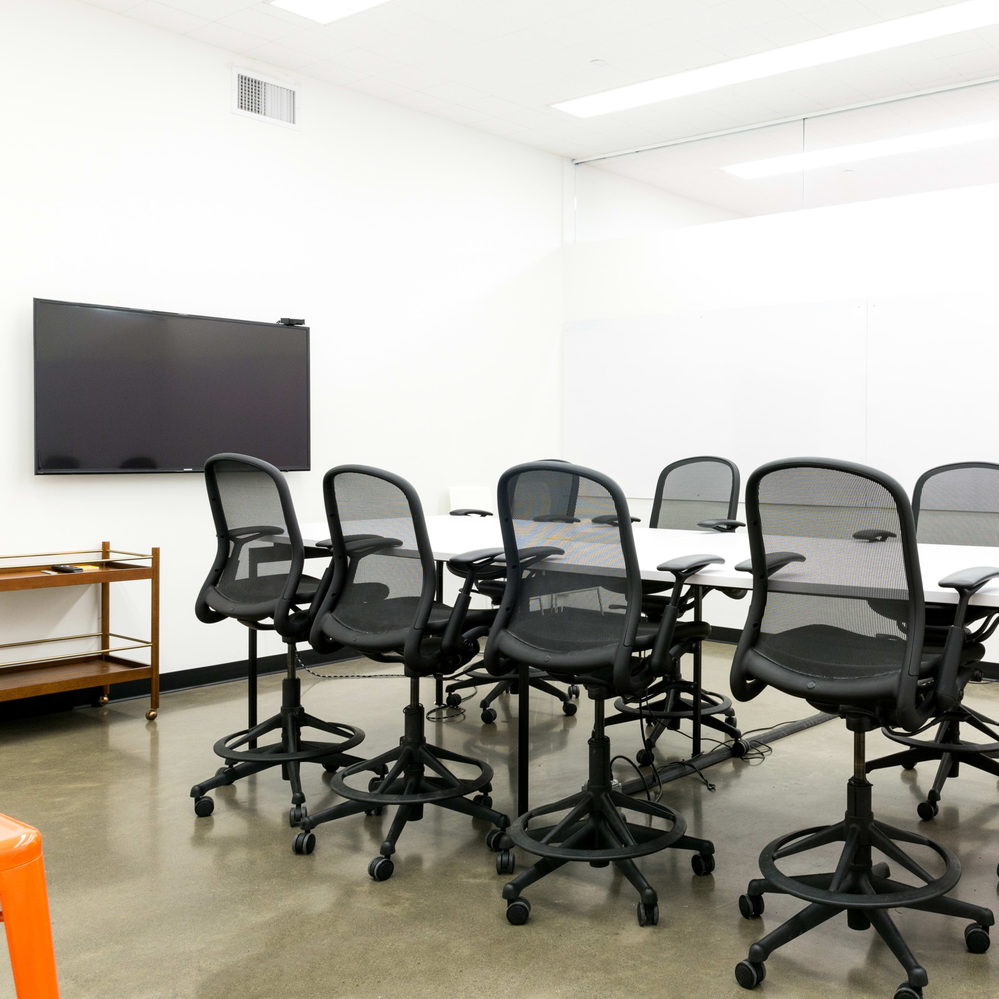 A conference room features a long table surrounded by adjustable chairs, with a television mounted on the wall and two orange stools in the corner.