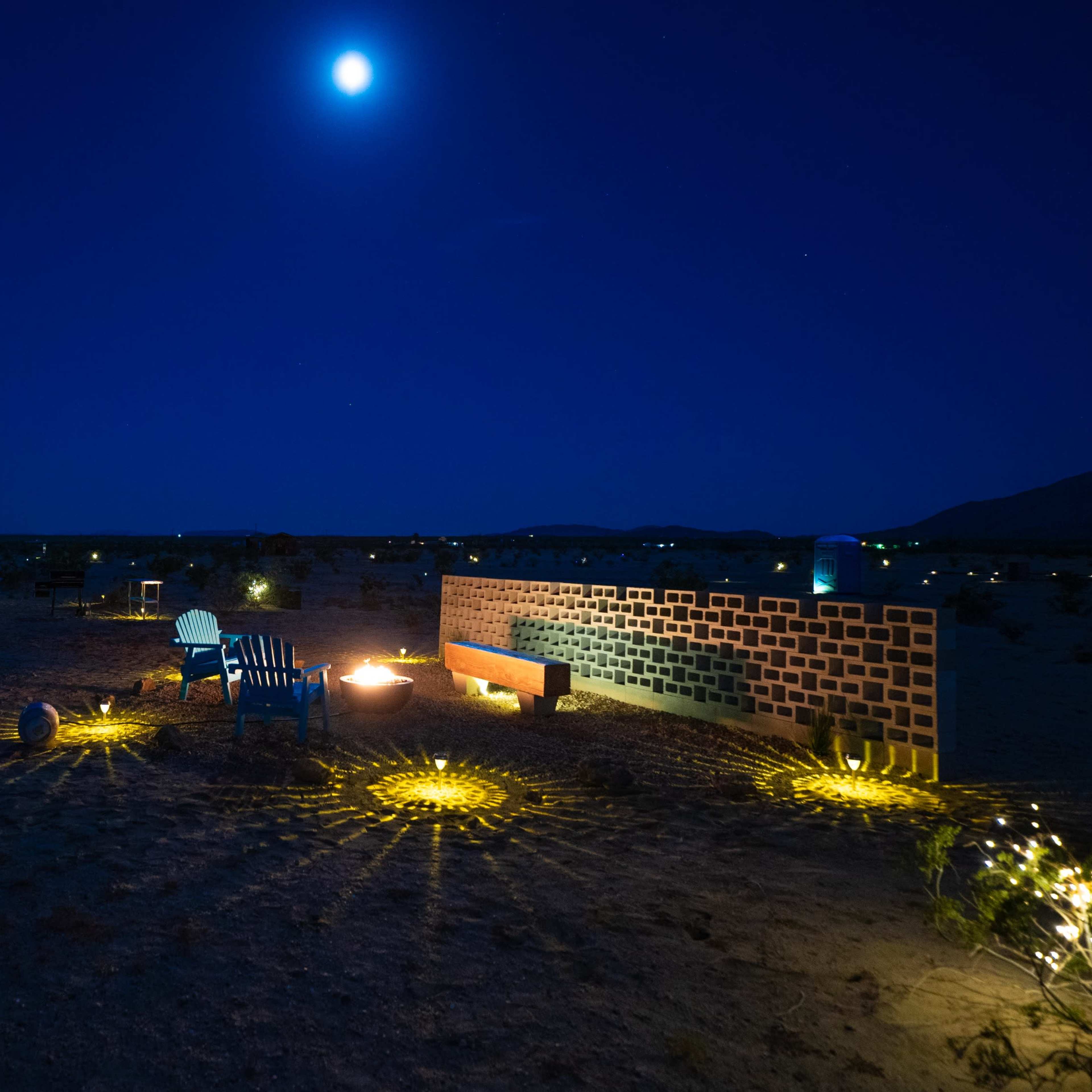 A small gathering area with colorful chairs and a brick wall is illuminated under a bright moon in a desert landscape.