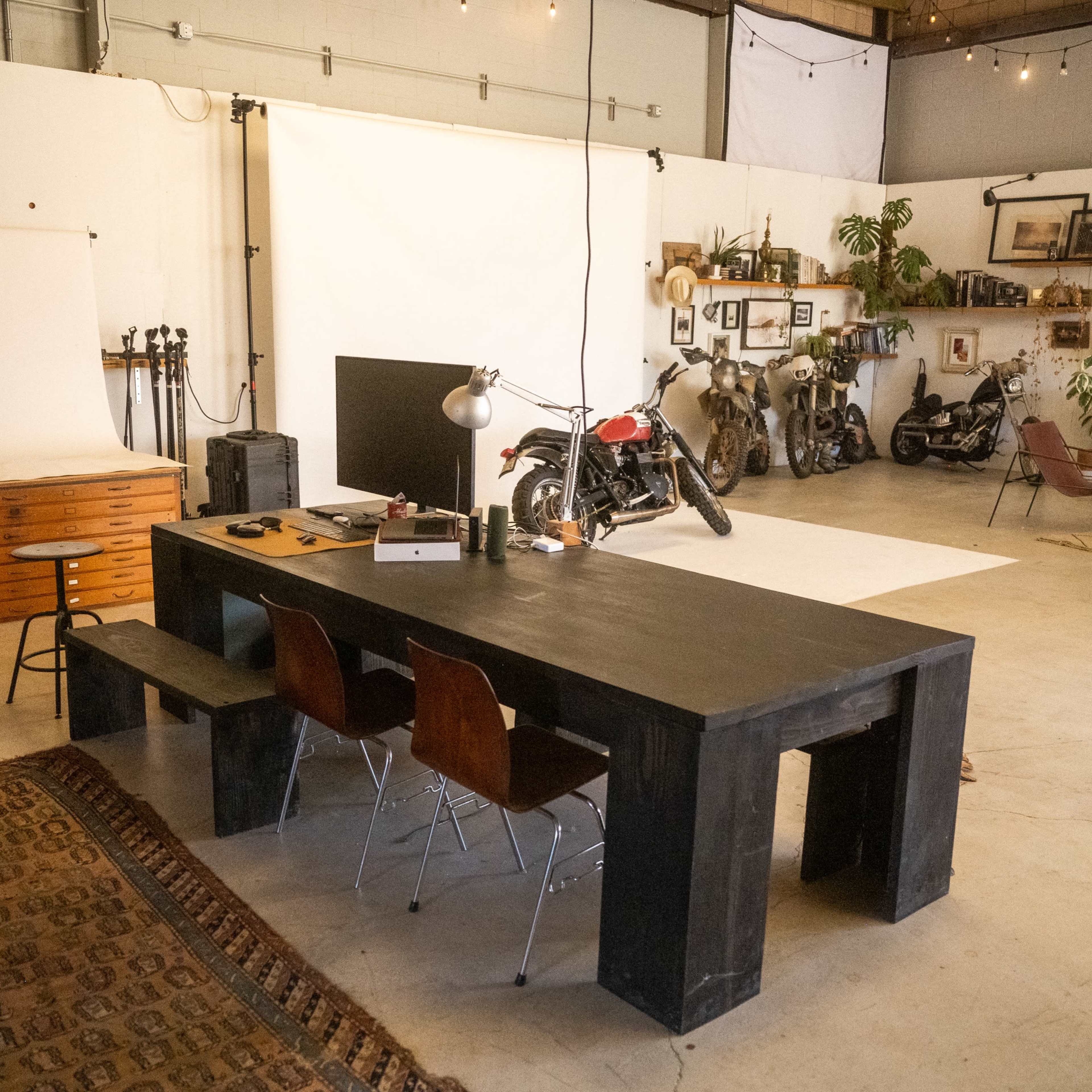 A spacious studio with a large black table, wooden chairs, a photography backdrop, vintage motorcycles, and various framed pictures on the walls.
