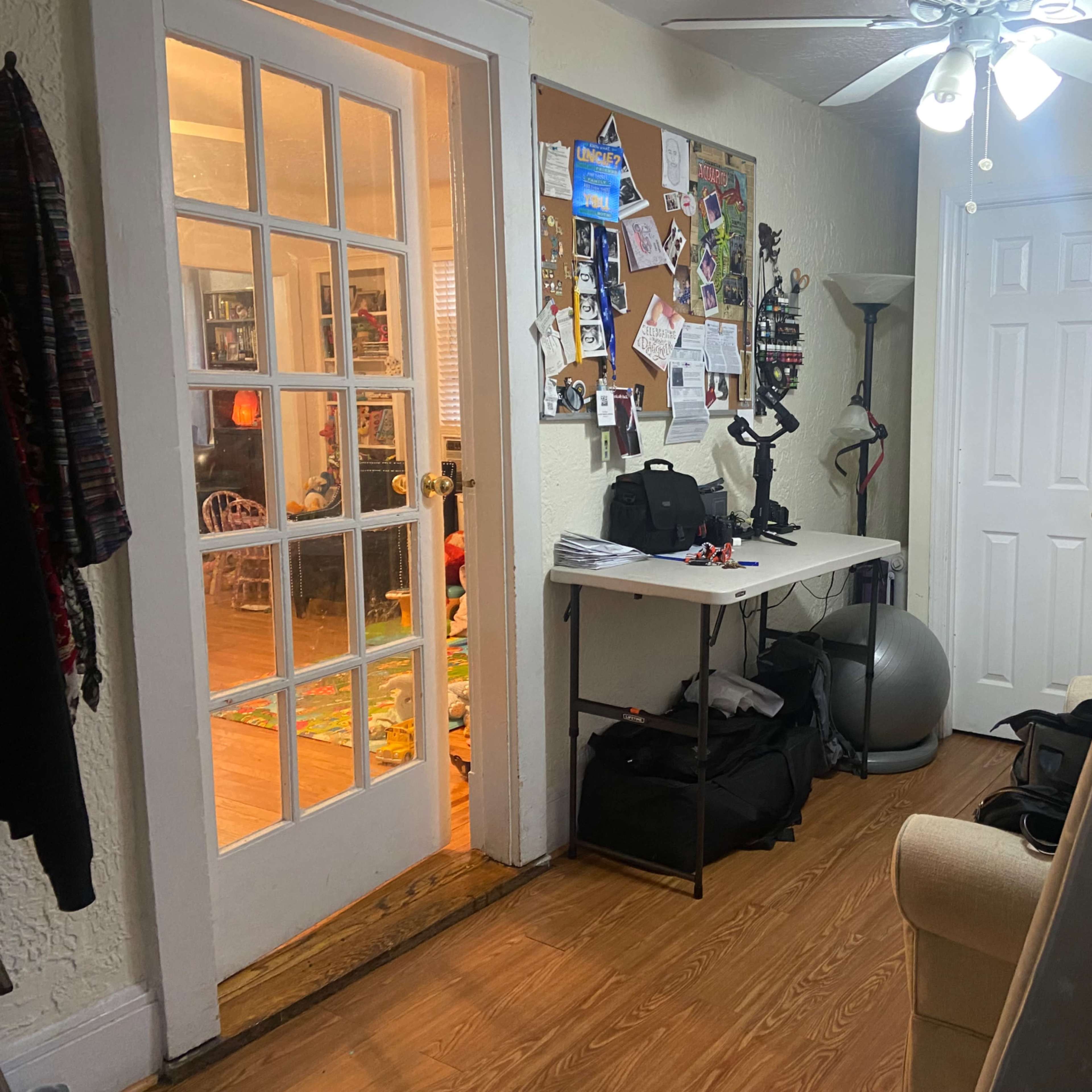 A hallway leads to a room with a bulletin board and various furniture items, including a table and exercise equipment.