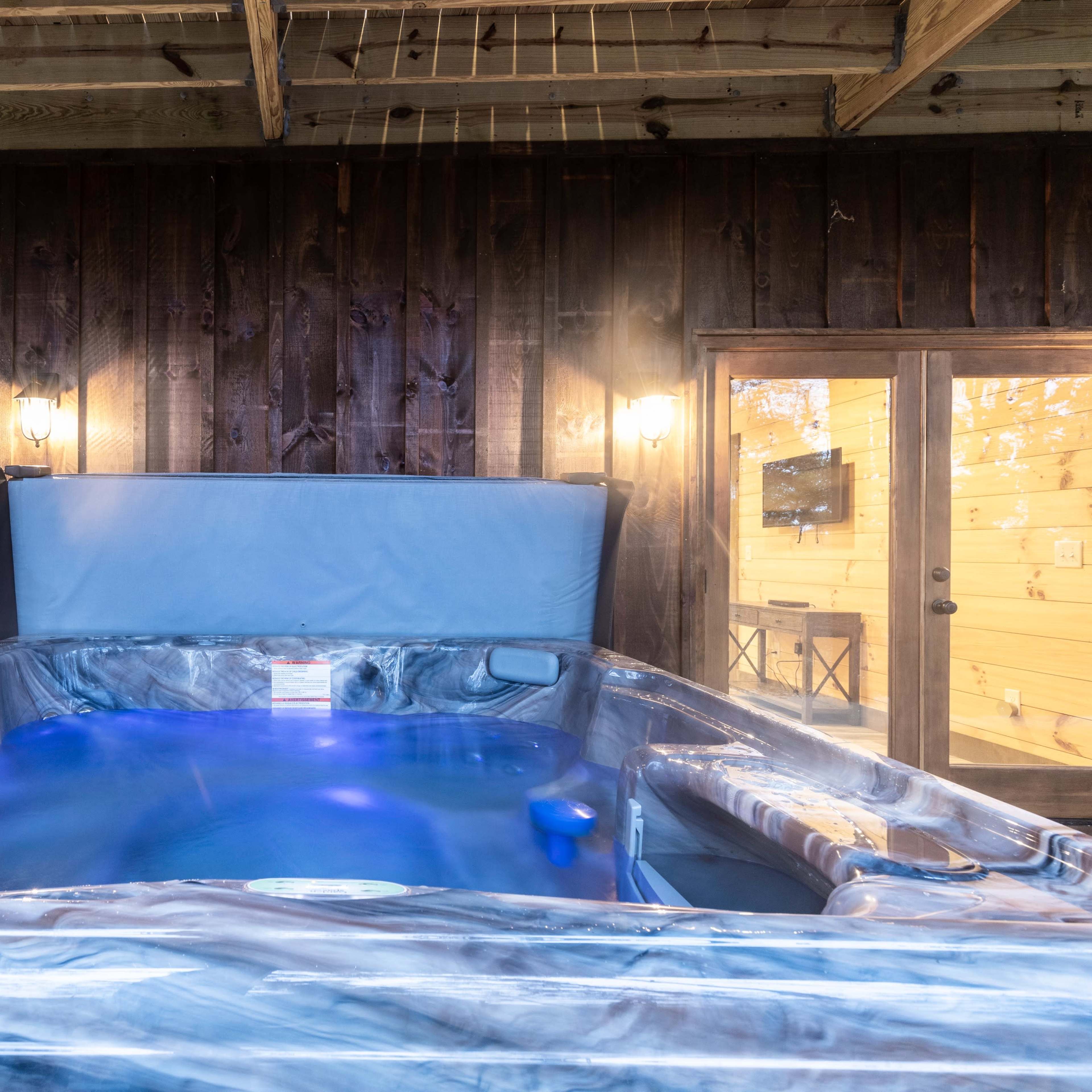 A hot tub with blue lighting is positioned under a wooden porch, adjacent to glass doors leading to a cabin interior.
