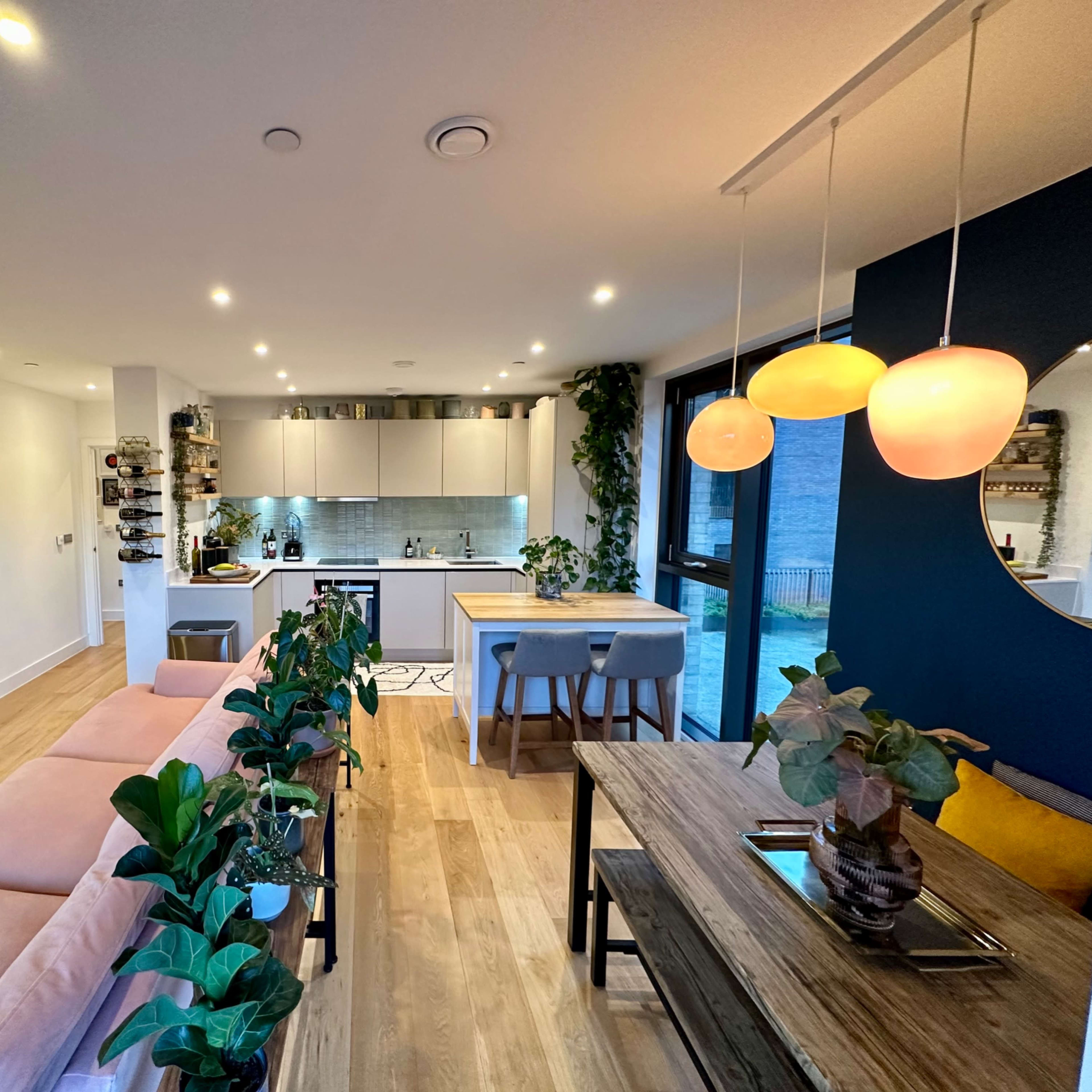 A modern living space with a light wood floor, a kitchen area in the background, and a dining table surrounded by plants.