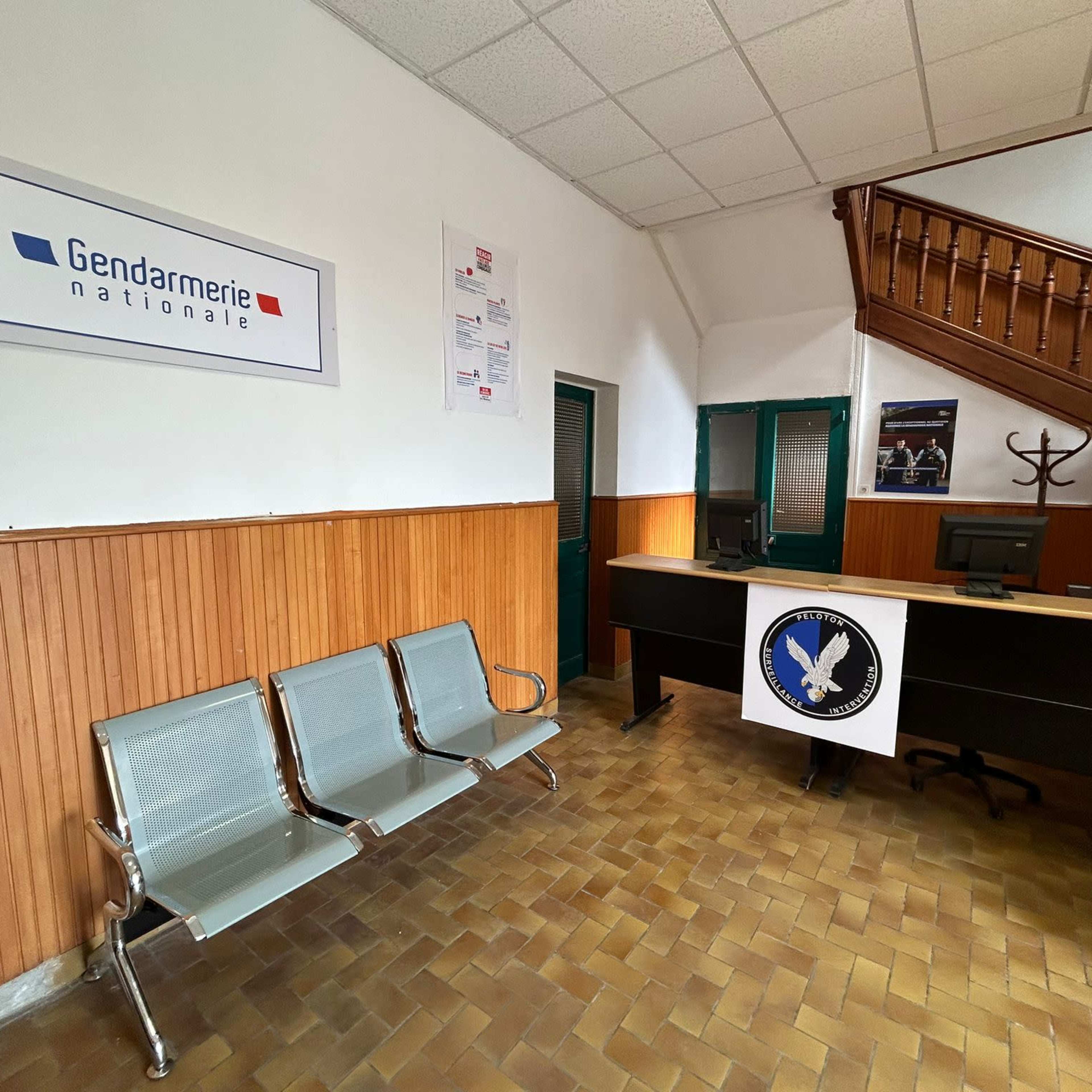 The interior of a gendarmerie station with metal benches, a reception desk displaying a logo, and wooden paneling on the walls.