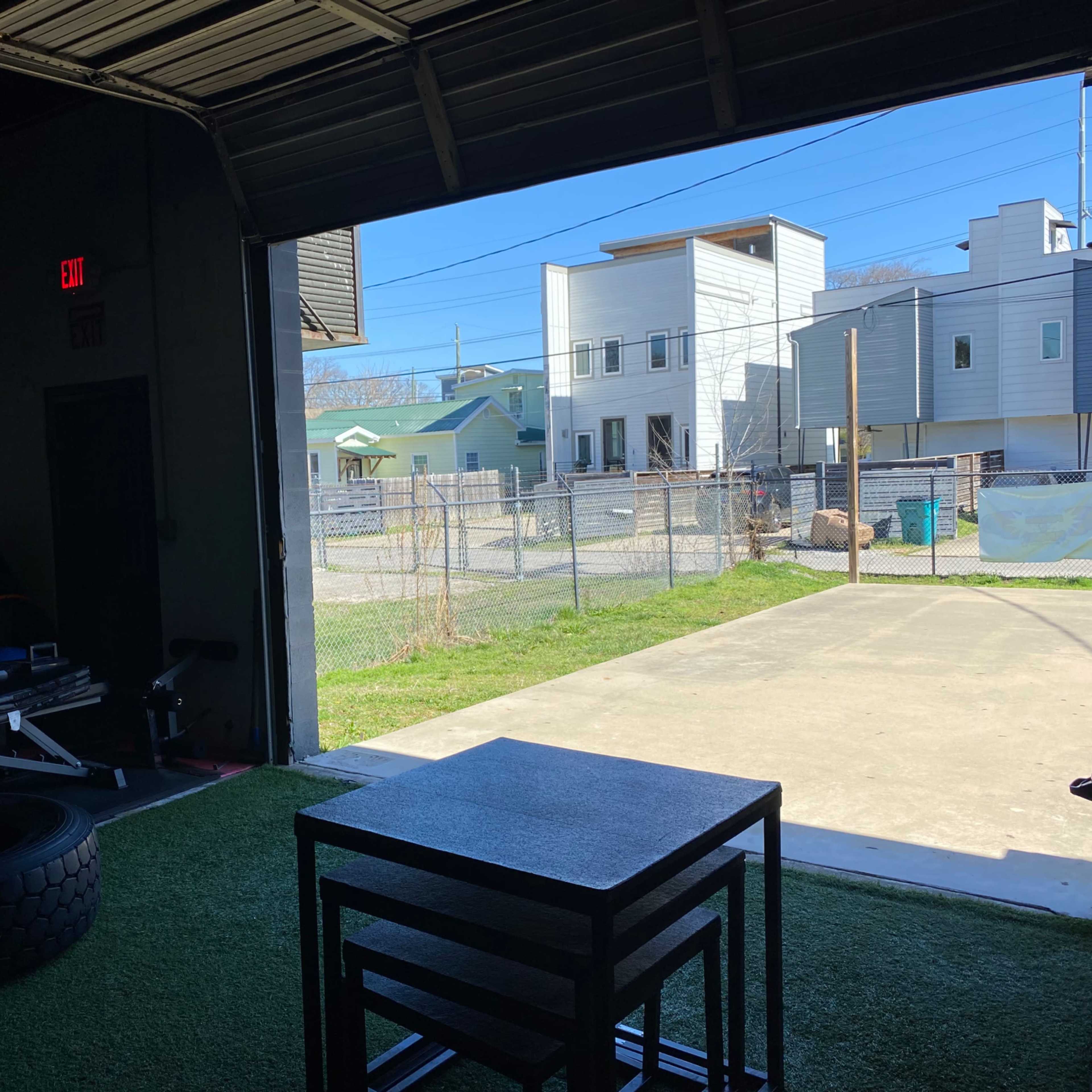 The interior of a gym with exercise equipment and a view of a fenced outdoor area and nearby buildings through an open garage door.