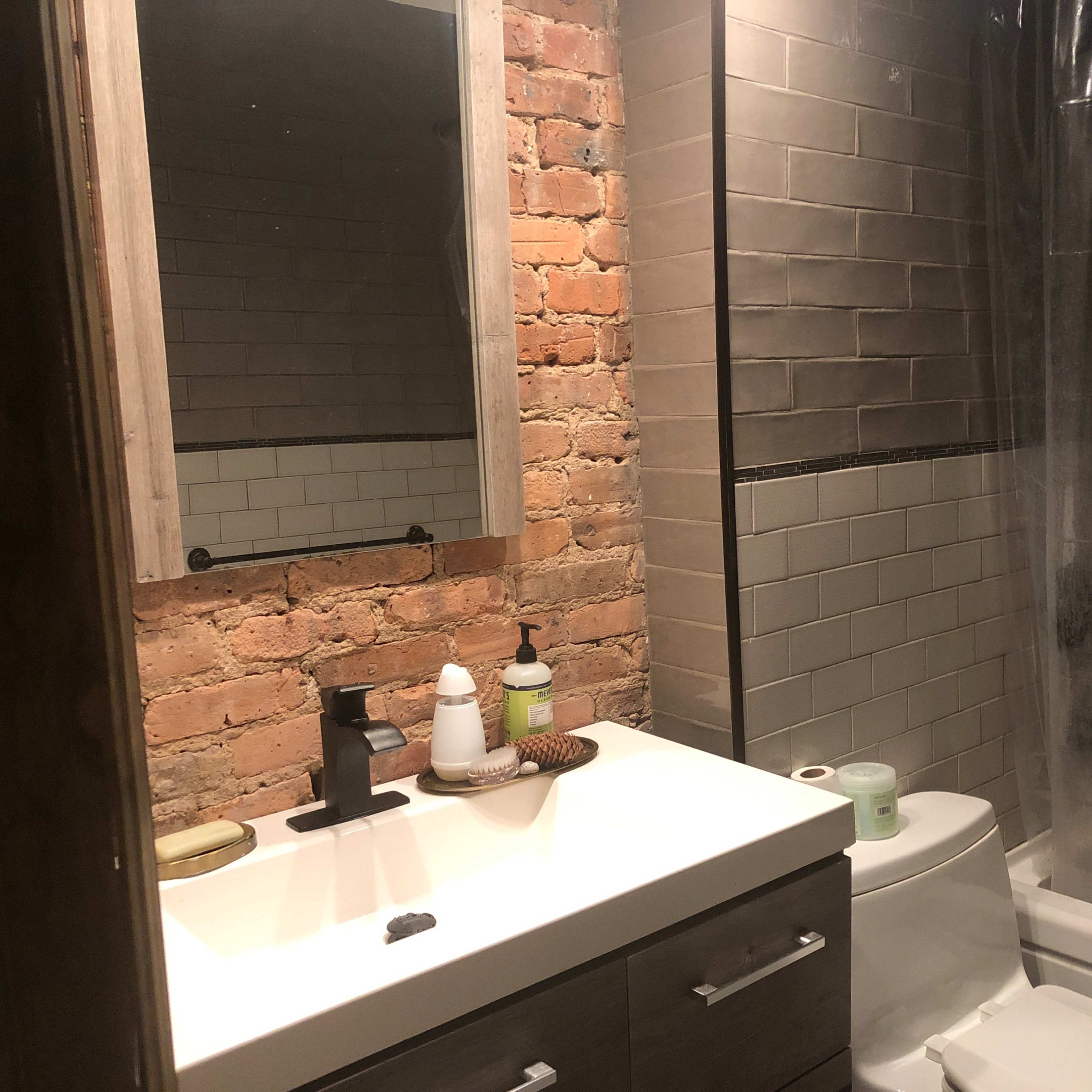 A bathroom featuring exposed brick walls, a modern sink with a mirror above it, a shower with a clear curtain, and a toilet placed next to the tub.