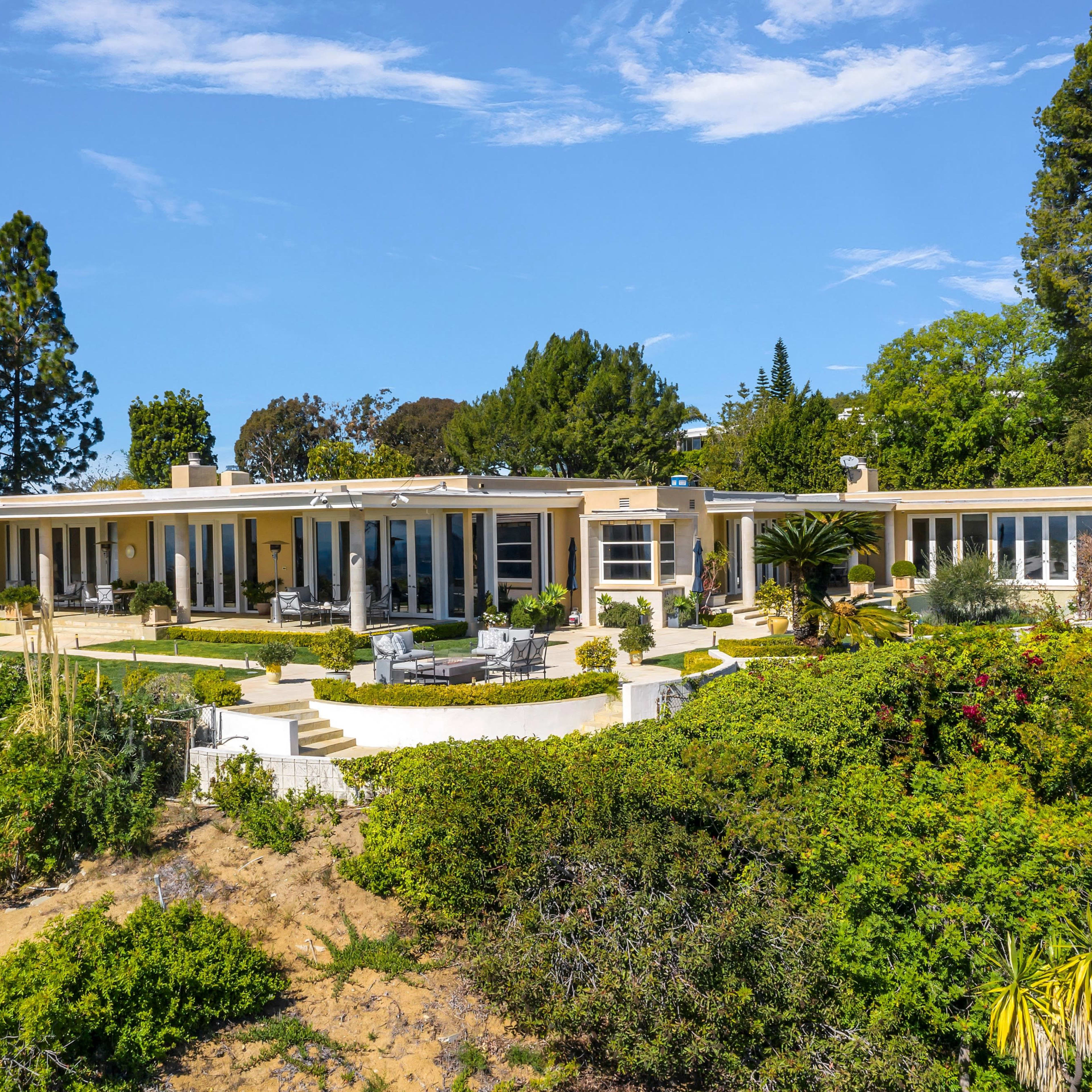 A single-story modern house with large windows is surrounded by lush landscaping and trees under a clear blue sky.