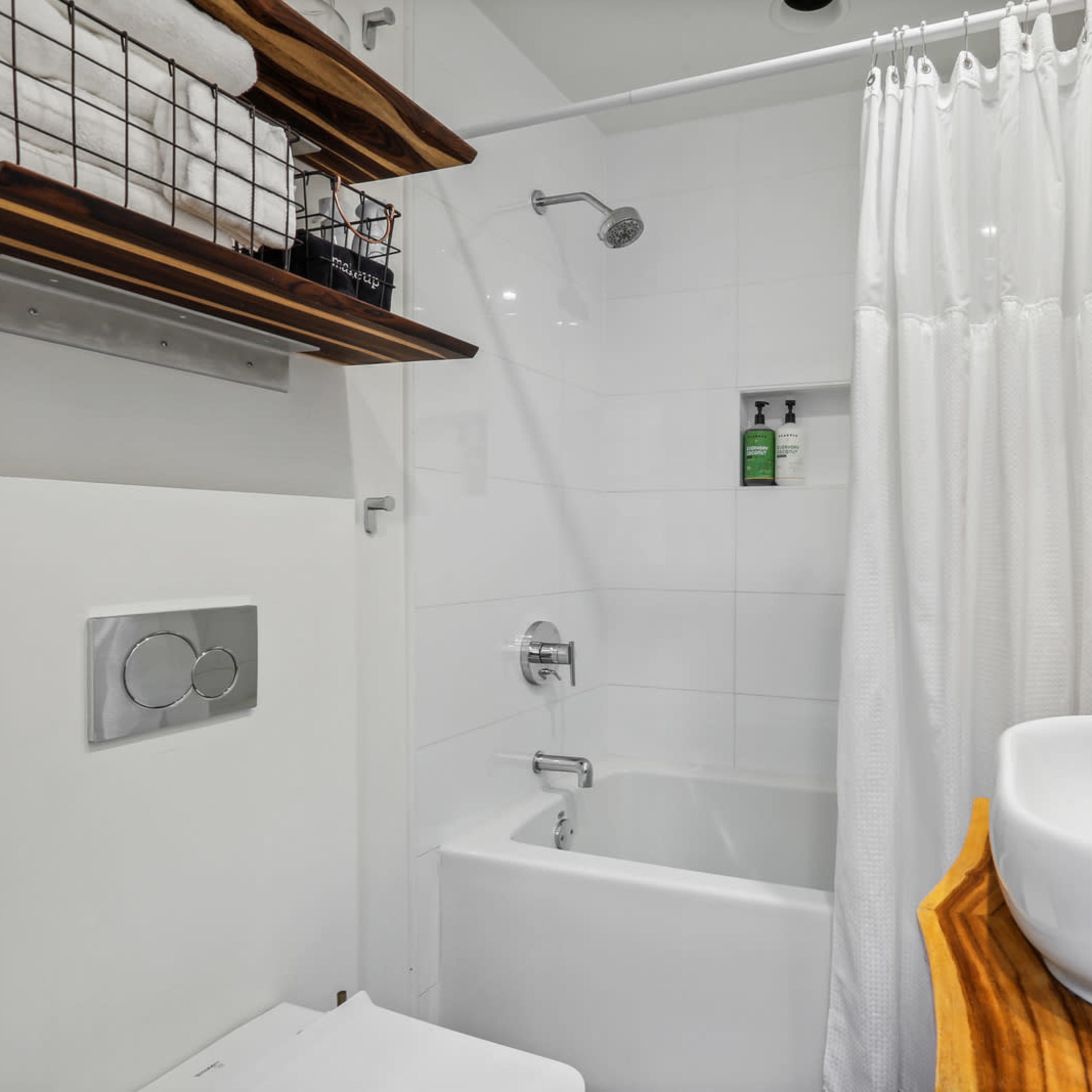 A modern bathroom featuring a bathtub with a shower, a sink on a wooden countertop, and neatly arranged towels on shelves.