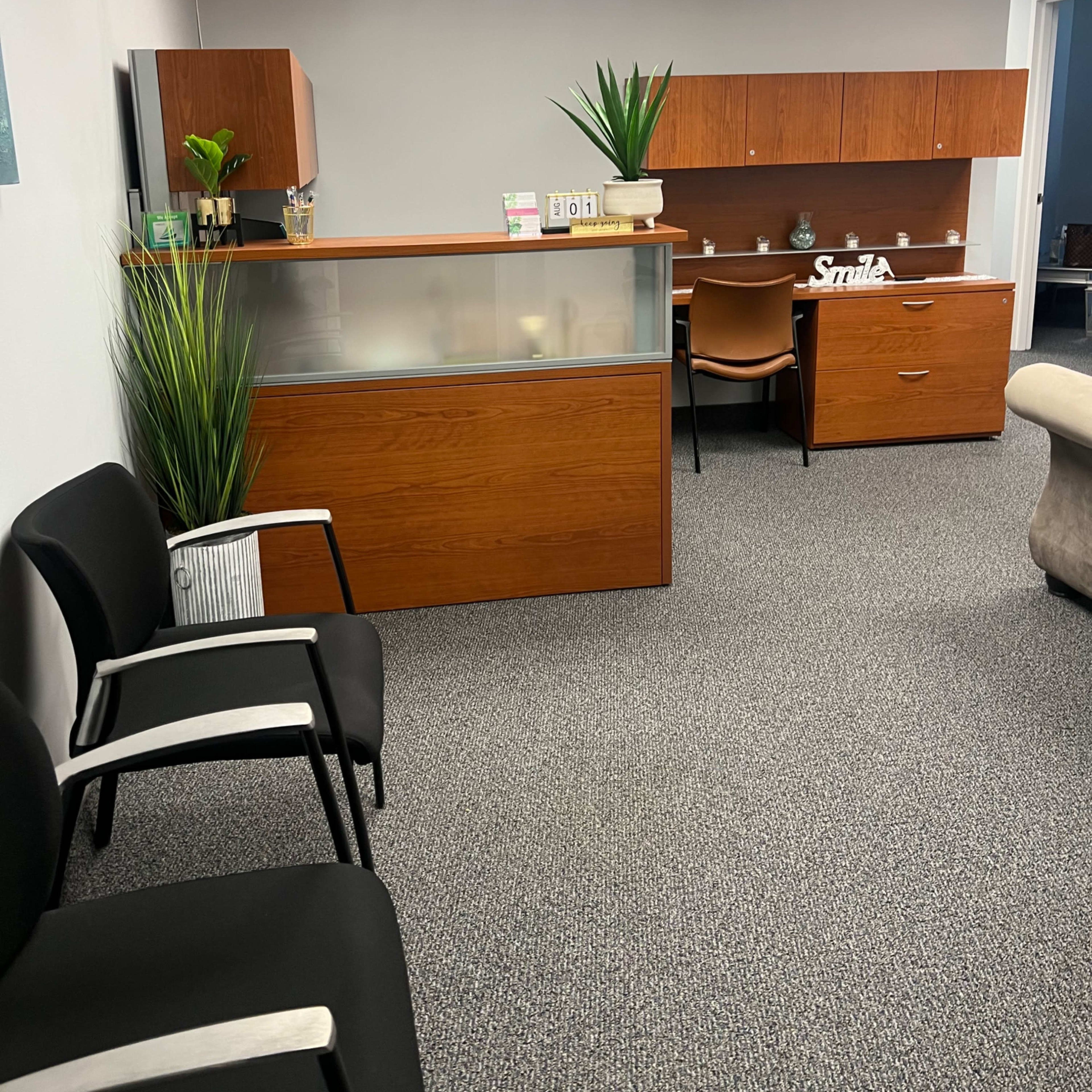 A reception area with wooden furniture, including a front desk and an office chair, surrounded by potted plants and carpeted flooring.
