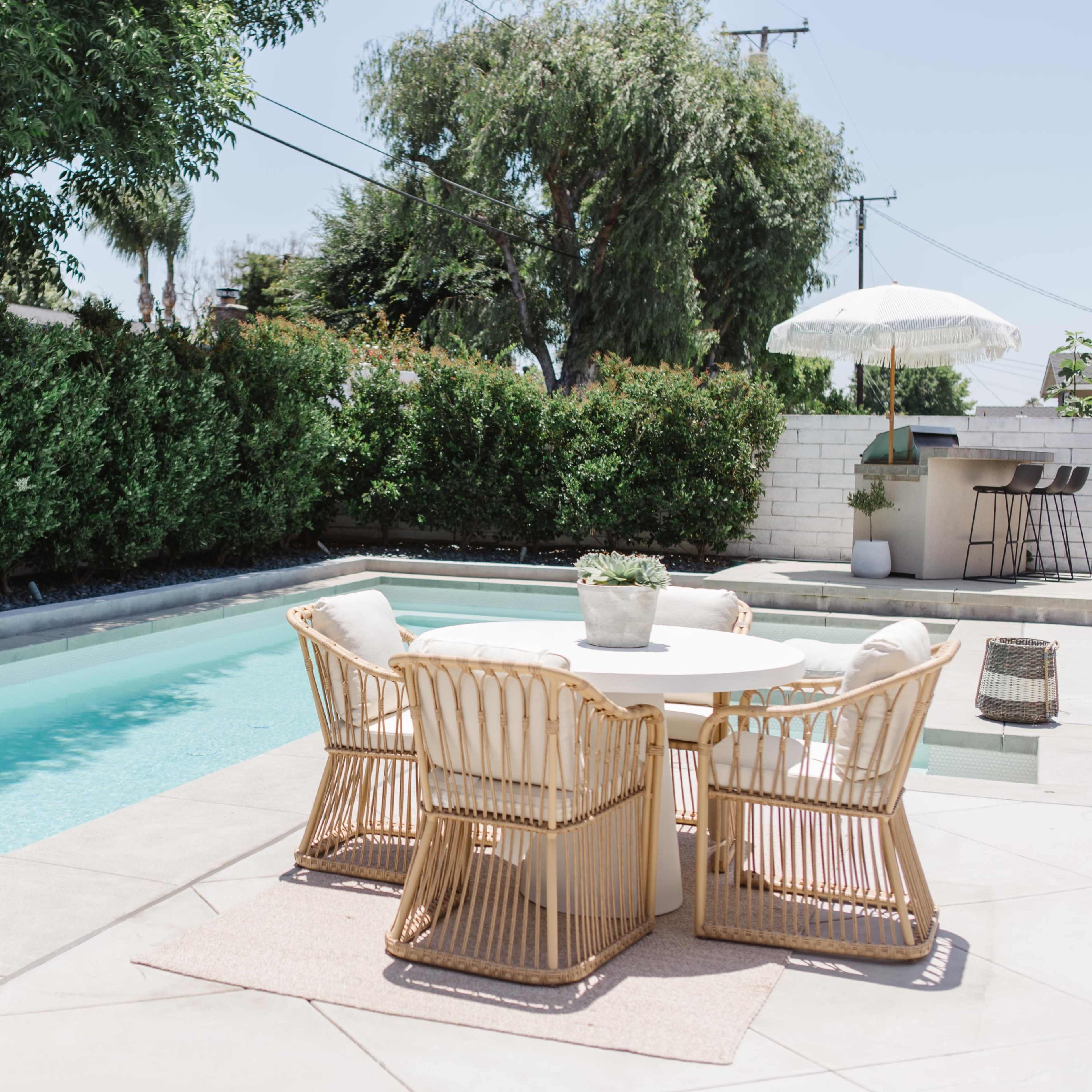 A round table with four wicker chairs sits beside a clear swimming pool, surrounded by lush greenery and a shaded area with a patio umbrella.