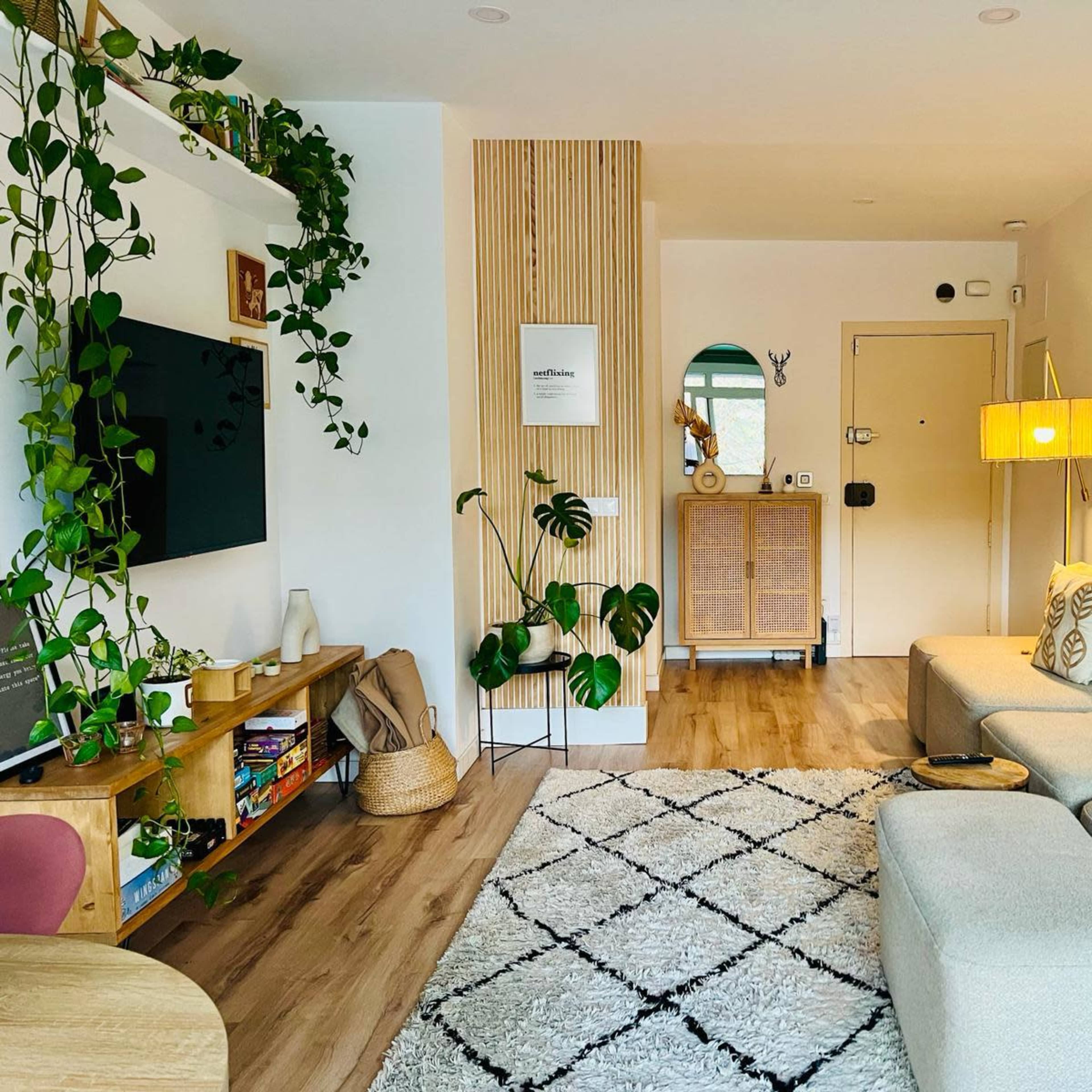 A modern living room with a large sectional sofa, a wooden TV stand, and indoor plants, leading to an entryway with a decorative mirror and a light-colored cabinet.