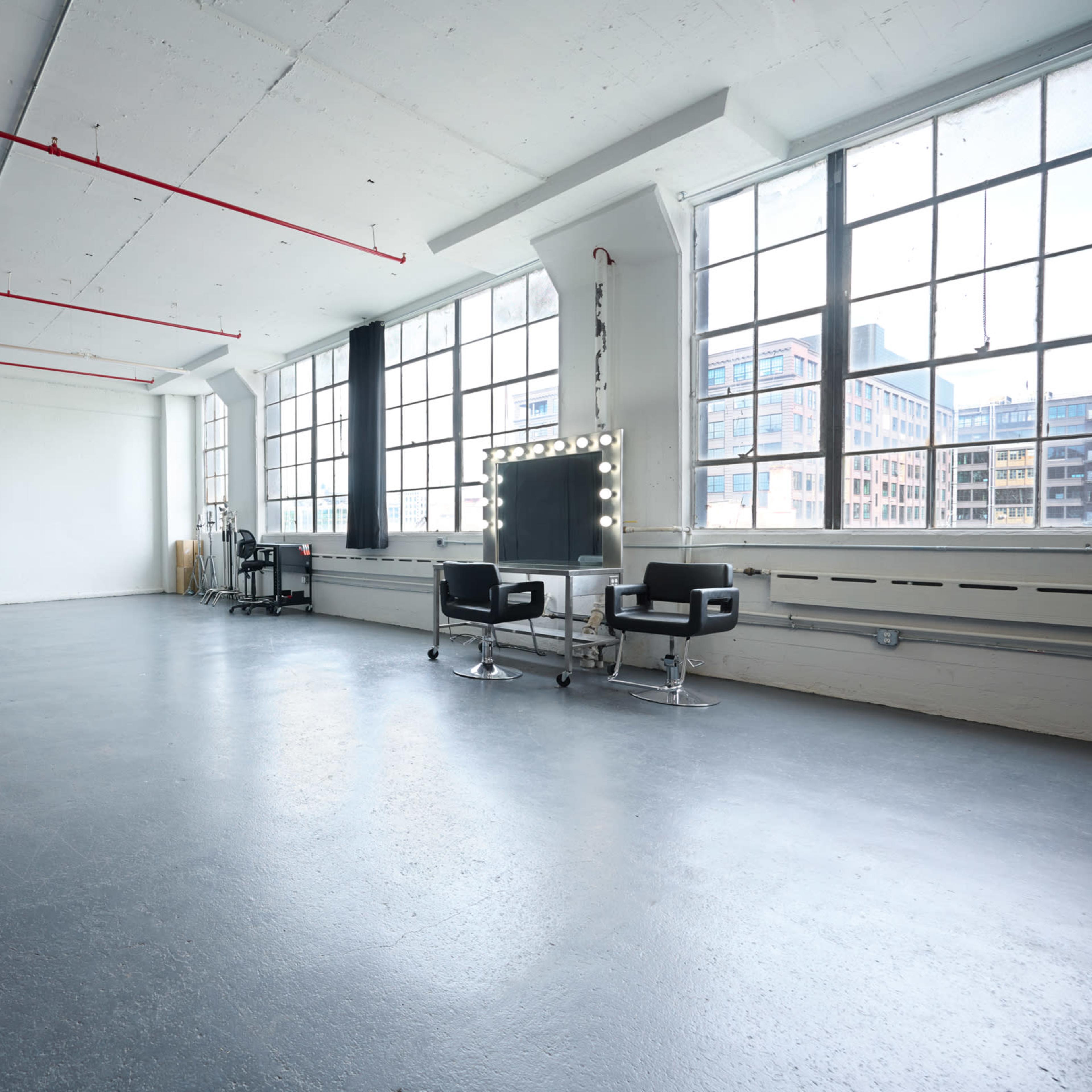 A spacious, well-lit studio with large windows, black curtains on one side, and two styling chairs with a mirror and lights at the front.