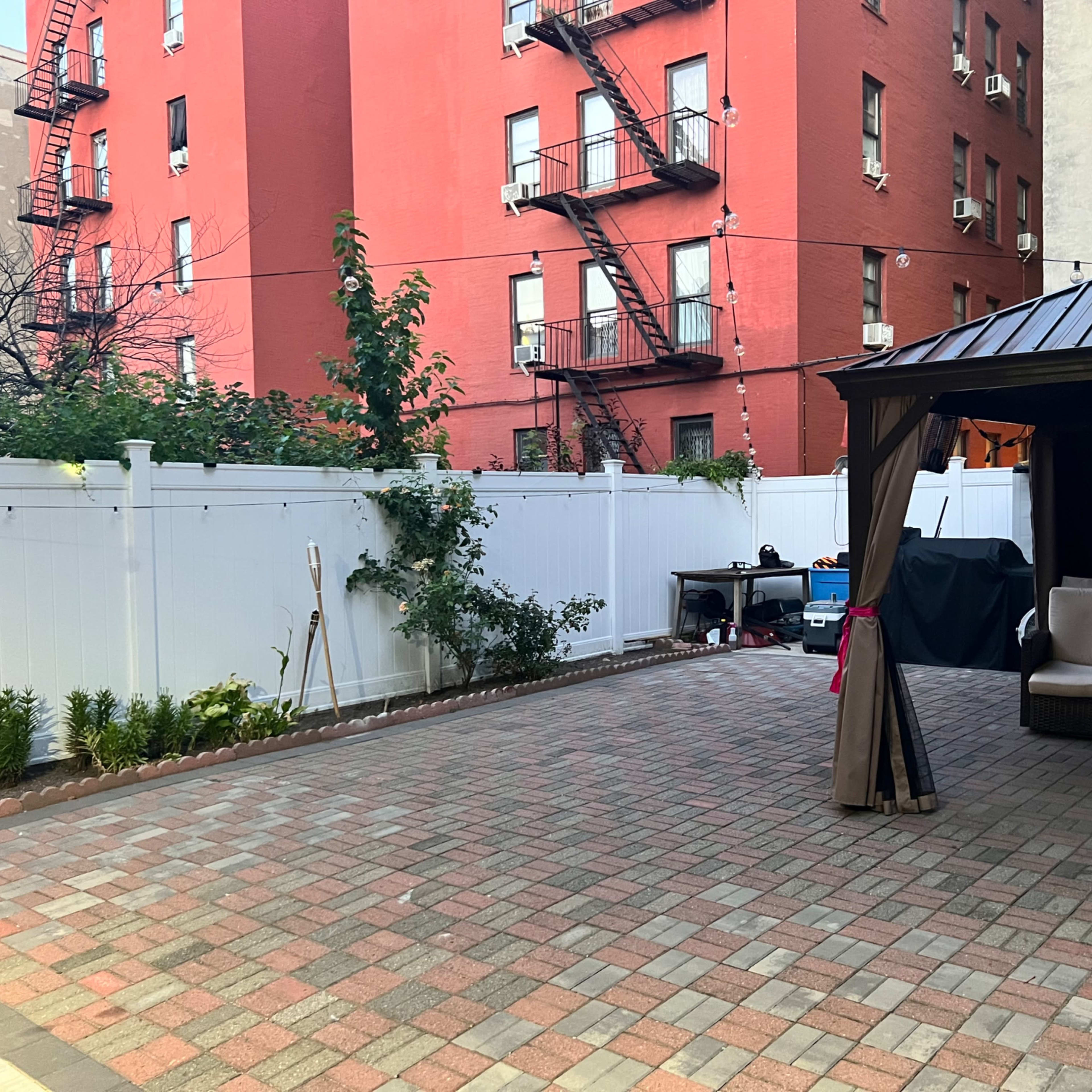 A paved backyard area with a gazebo and seating, surrounded by a white fence and red brick buildings in the background.