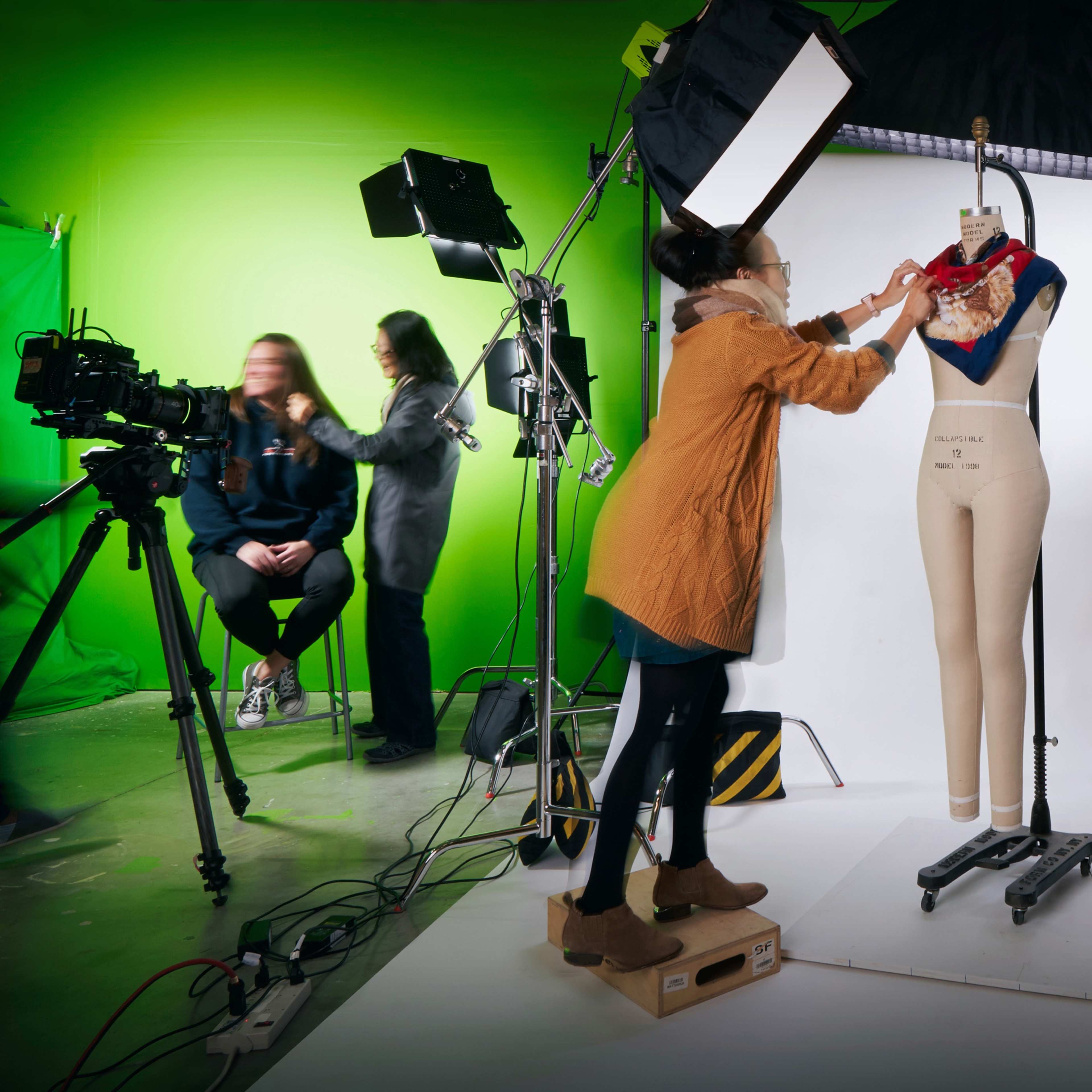 A person adjusts a scarf on a mannequin in a photography studio with green and white backgrounds and multiple lighting setups.