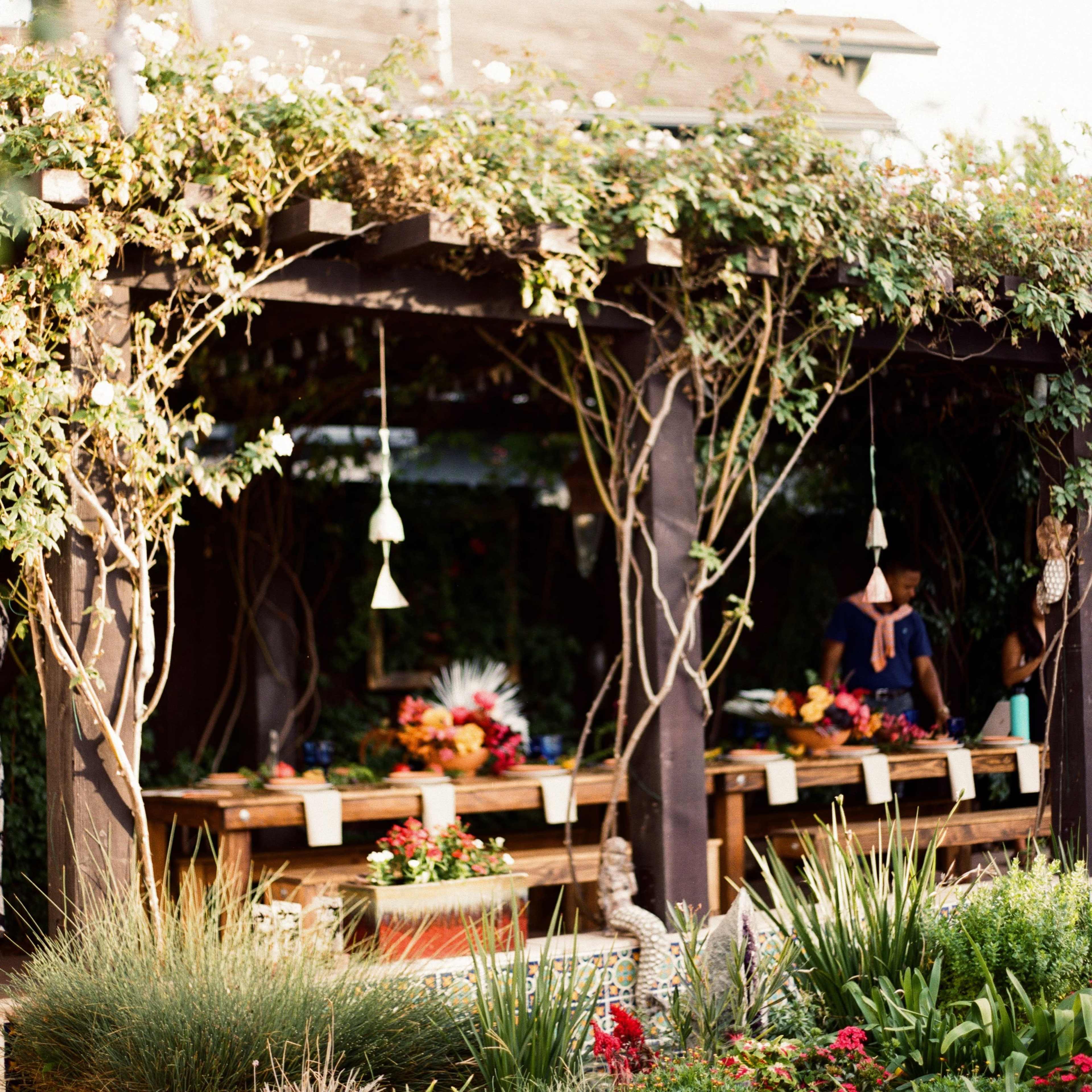 A wooden pergola covered in vines supports hanging decorations and frames a dining area adorned with floral arrangements.