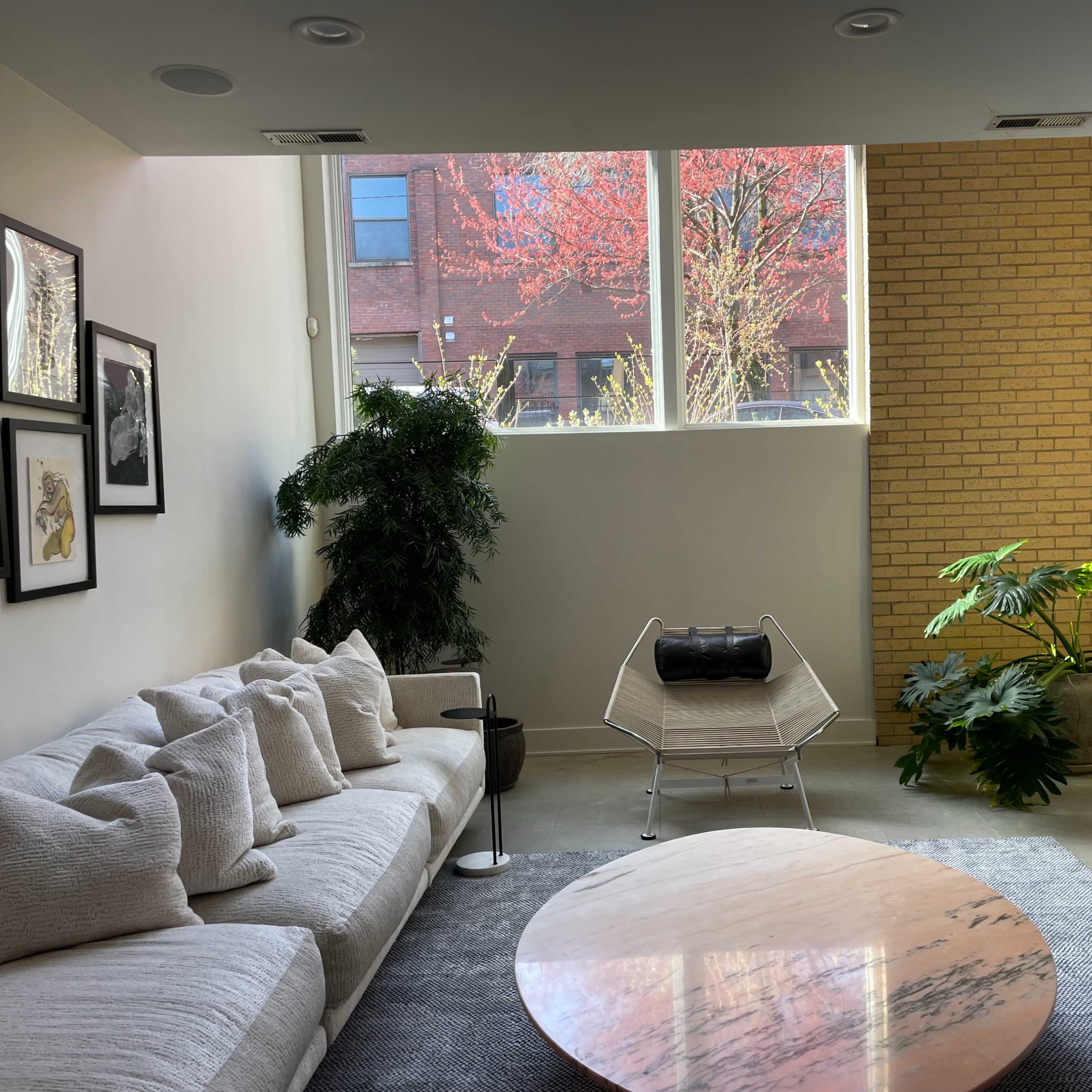 A modern living room featuring a white couch, a round marble coffee table, a potted plant, and large windows overlooking a brick wall with a view of trees outside.