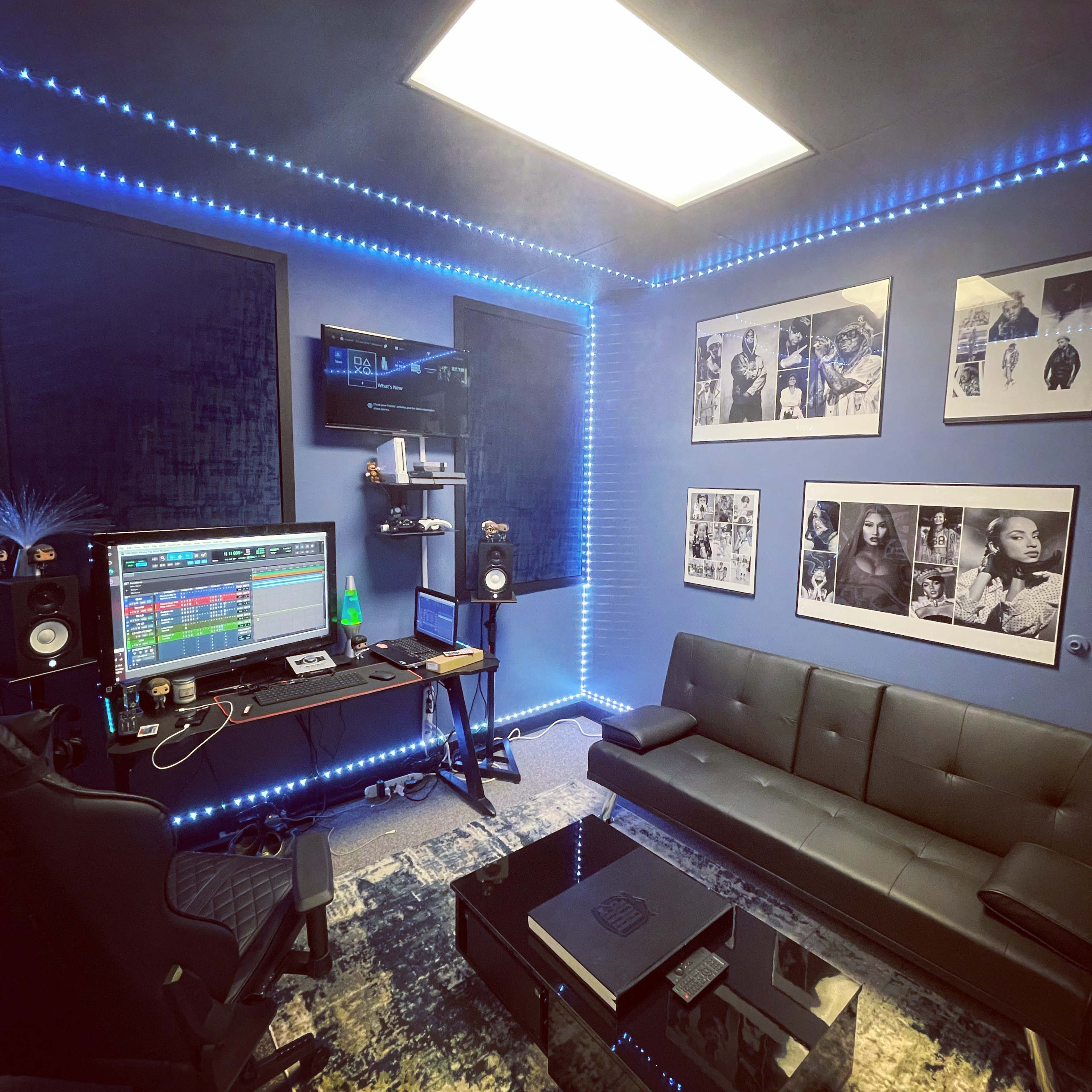 A music studio featuring a computer desk with audio equipment, a dark sofa, decorative wall art, and blue LED lighting.