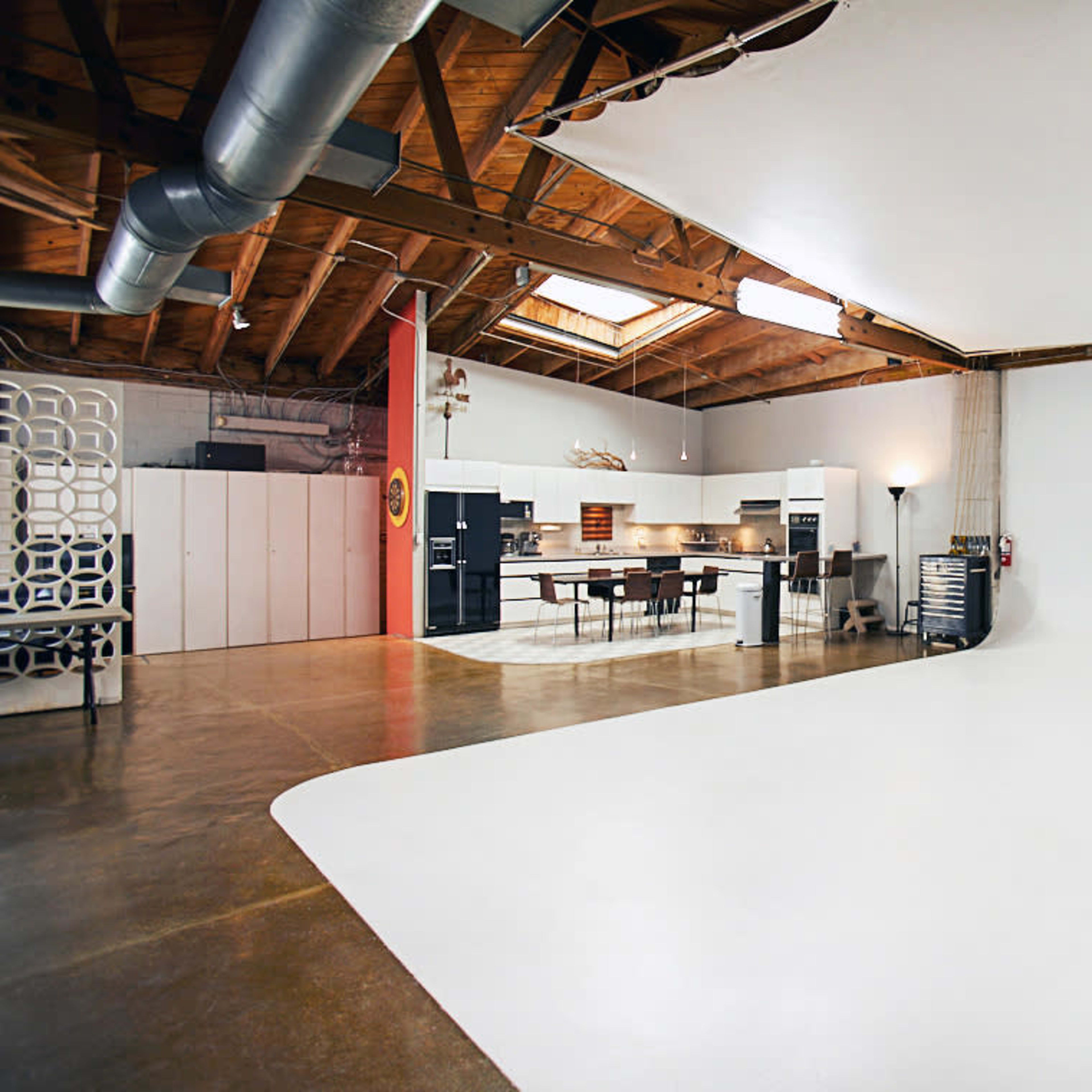 An industrial-style studio space with a large white backdrop, wooden beams, and a kitchen area in the background.