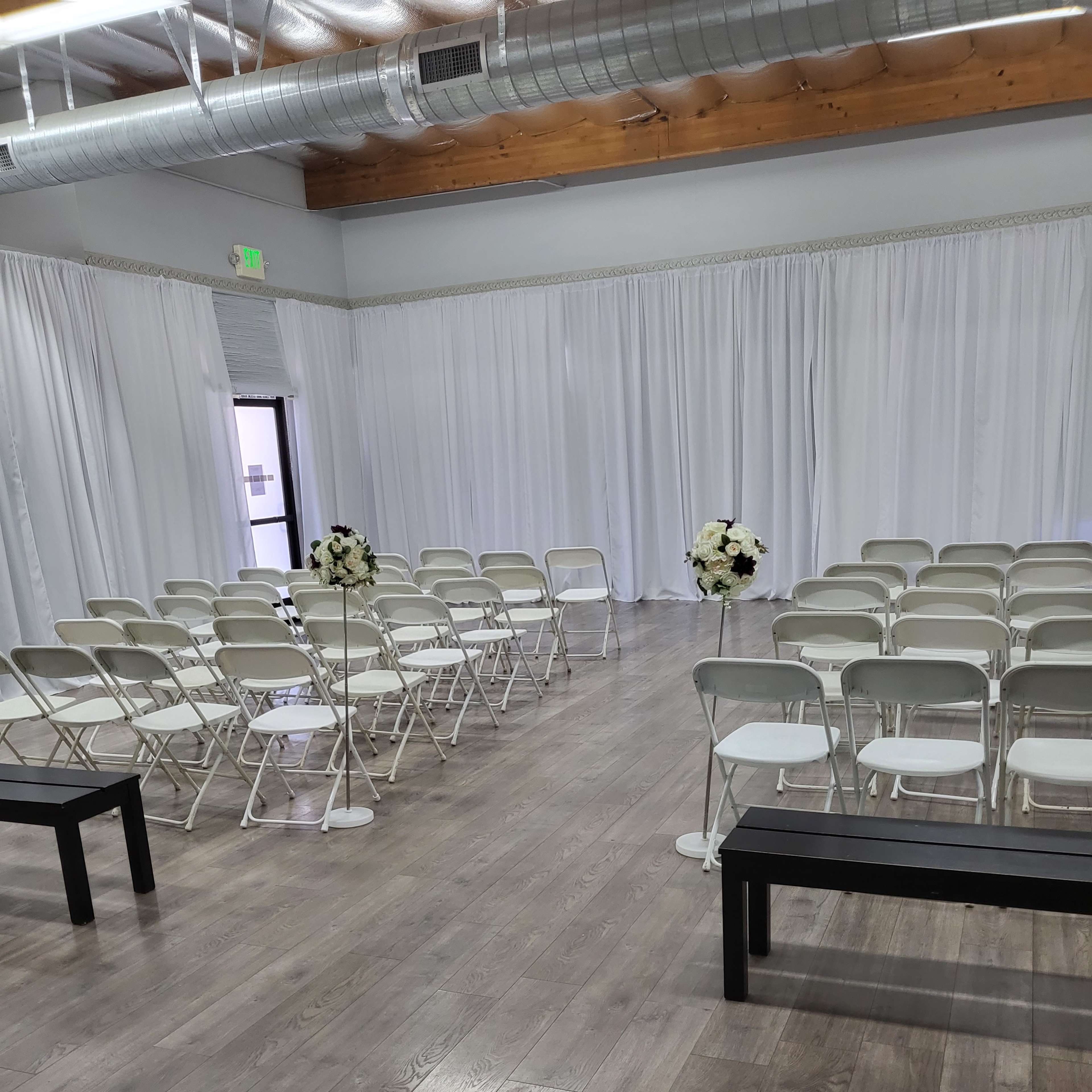 A spacious indoor venue set up for a ceremony, featuring rows of white folding chairs and flower arrangements on pedestals.