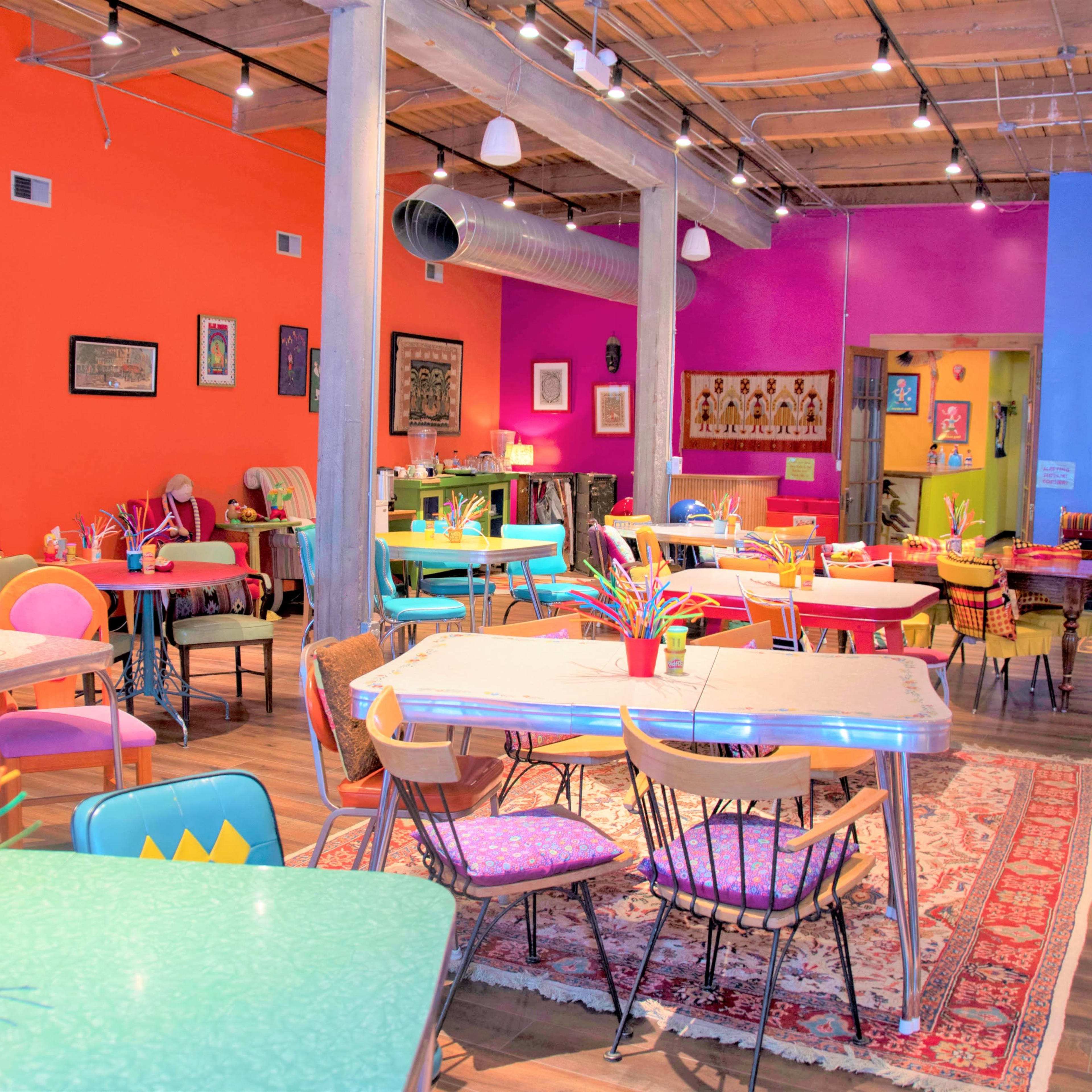 A vibrant cafe features colorful tables and chairs, craft supplies, and eclectic artwork on the walls.