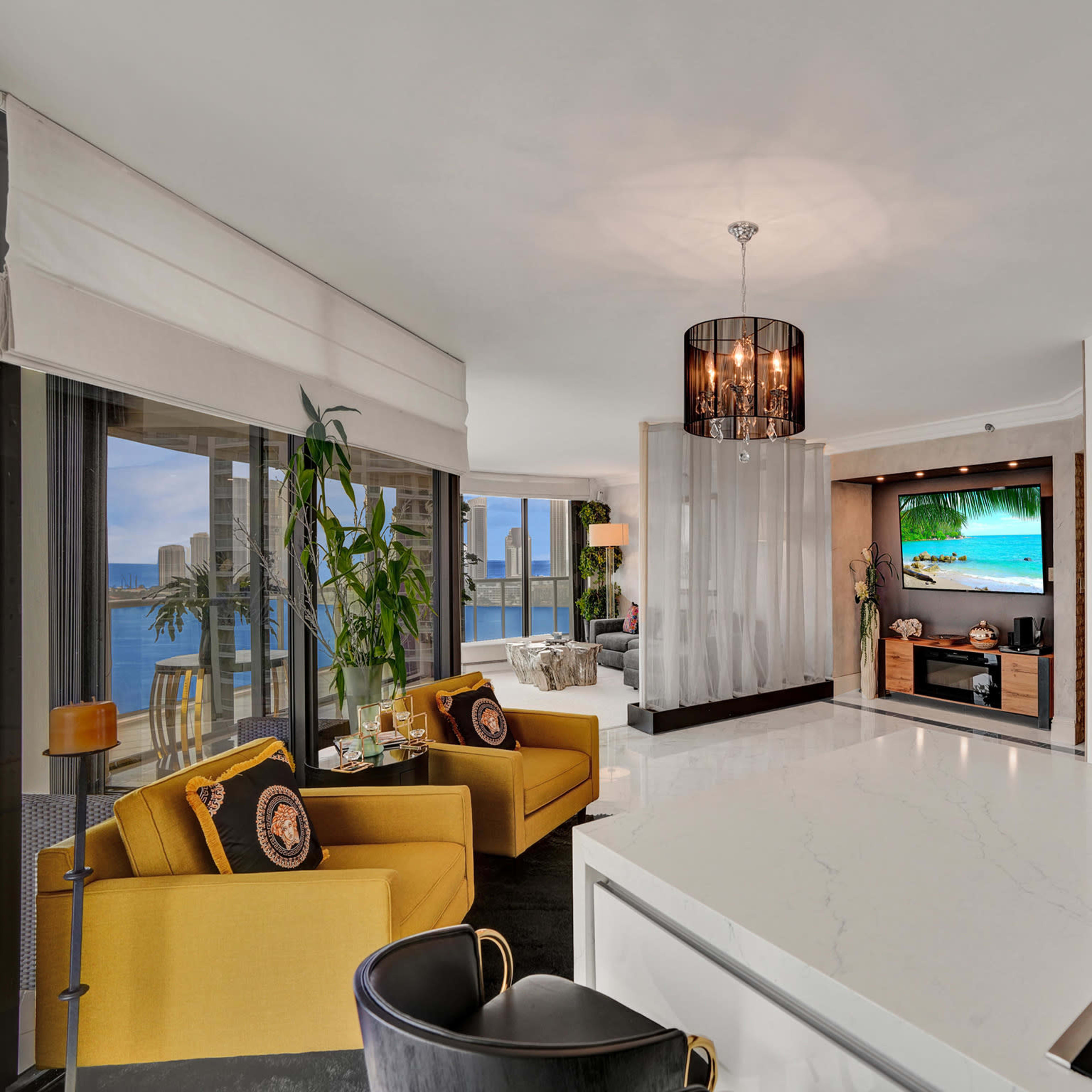 A contemporary living space featuring a kitchen island with a marble countertop, a seating area with yellow chairs, large windows offering a view of the sea, and a television mounted on a wall.