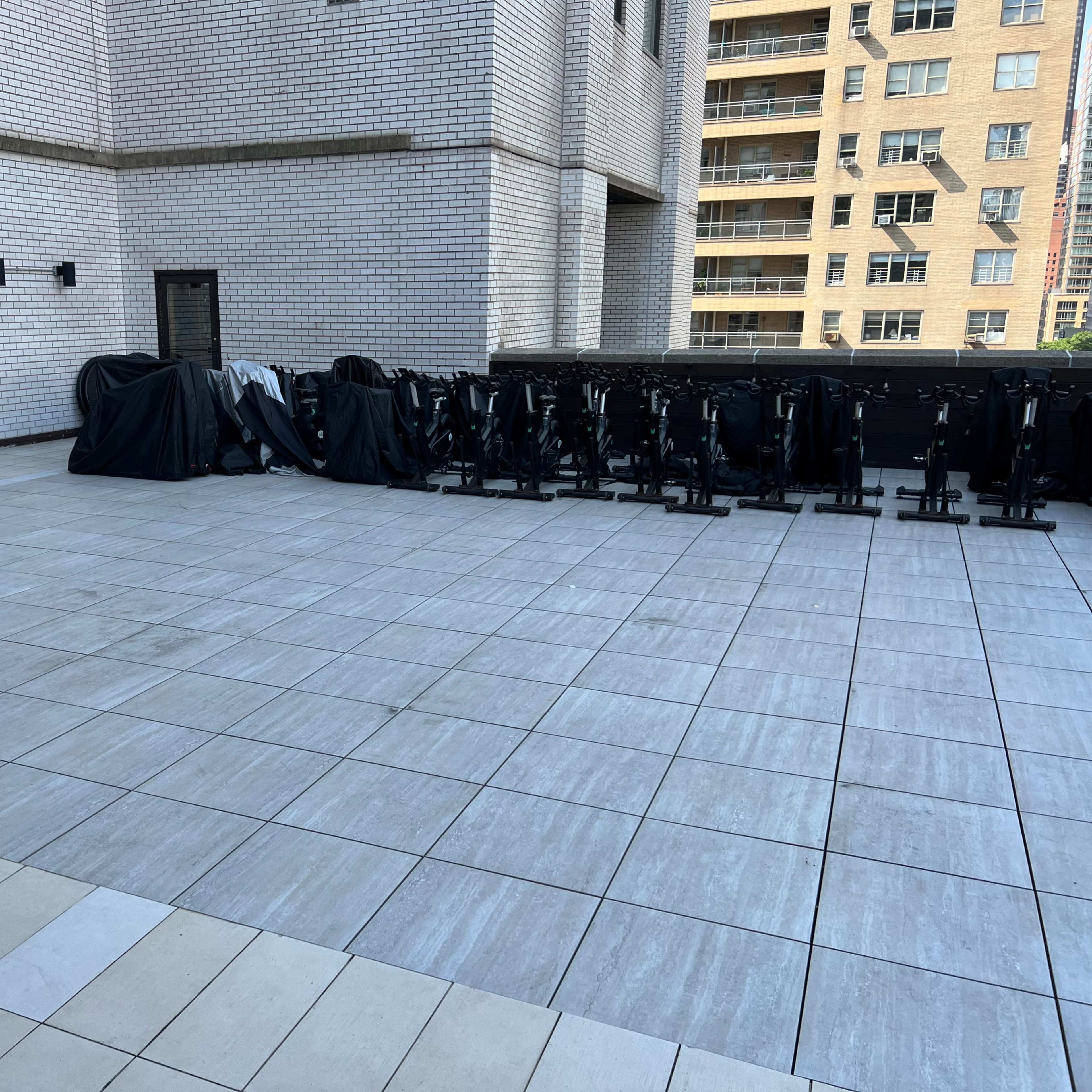 A rooftop terrace with gray tiled flooring, featuring a bar area and several stationary bicycles covered with black tarps against a wall.