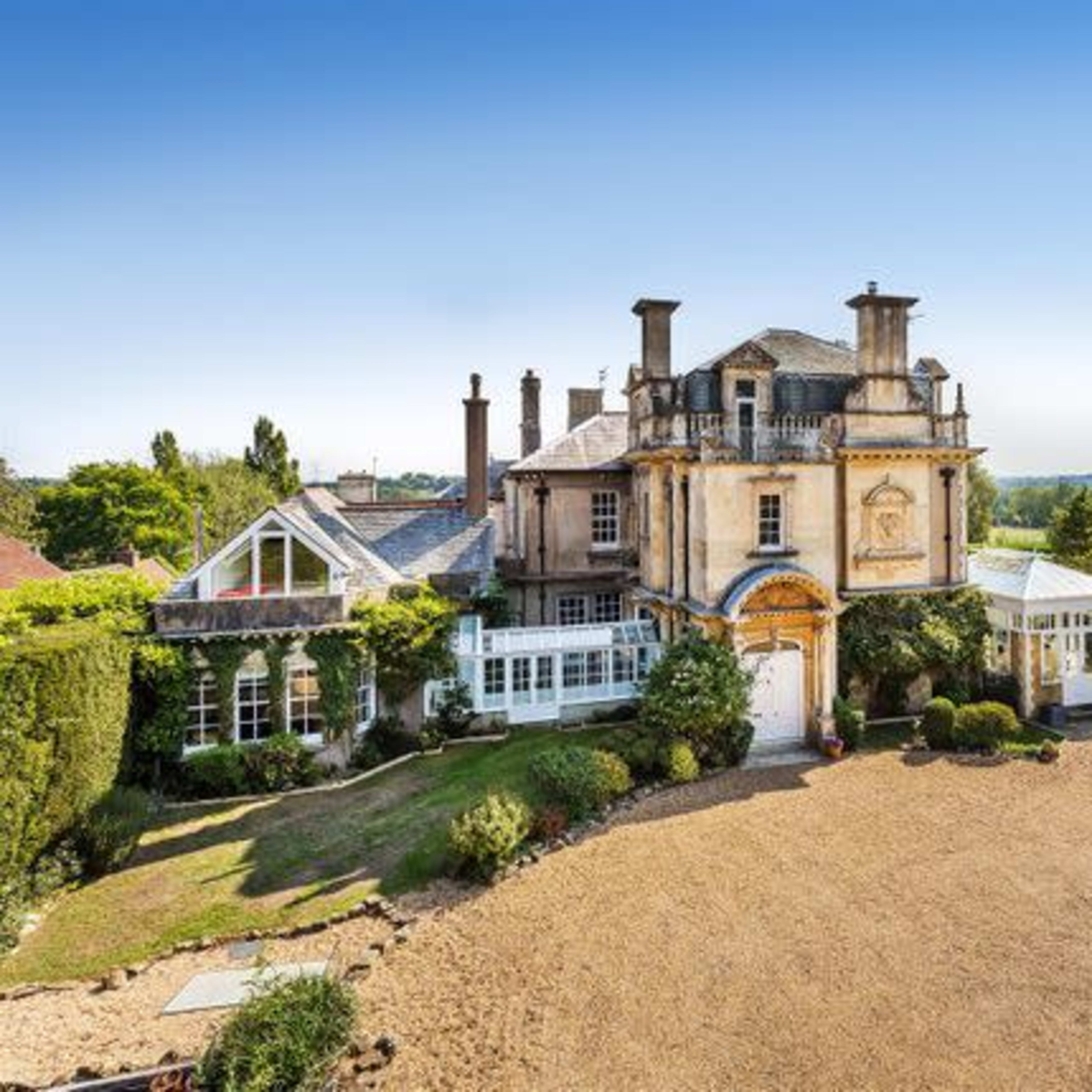 A large, elegant mansion surrounded by well-maintained gardens and trees under a clear blue sky.